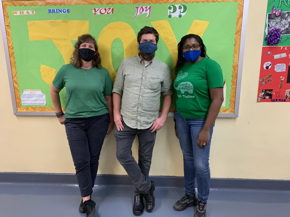 Three masked staff members posing in front of a Joy bulletin board