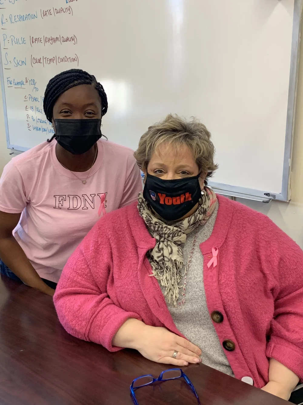 Two masked staff members in pink near a classroom whiteboard