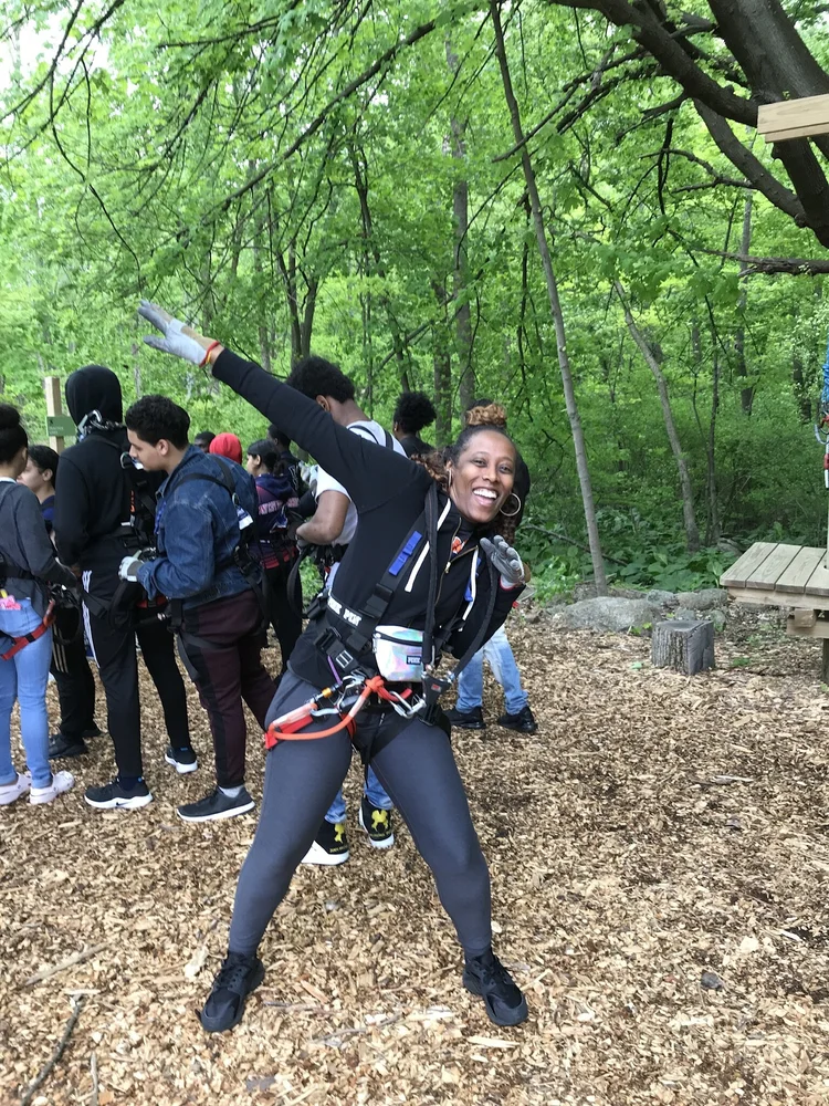 Adult wearing climbing harness posing cheerfully at outdoor ropes course with students