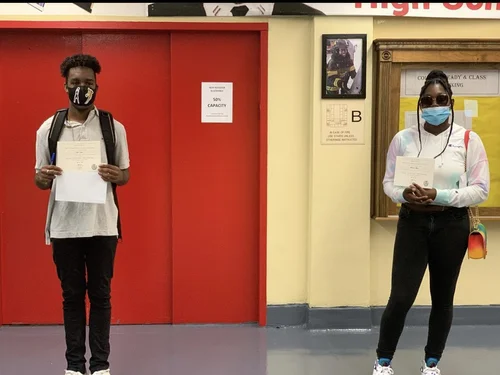 Two masked students holding certificates in school hallway