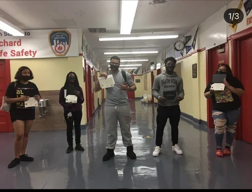Five masked students standing in school hallway holding papers