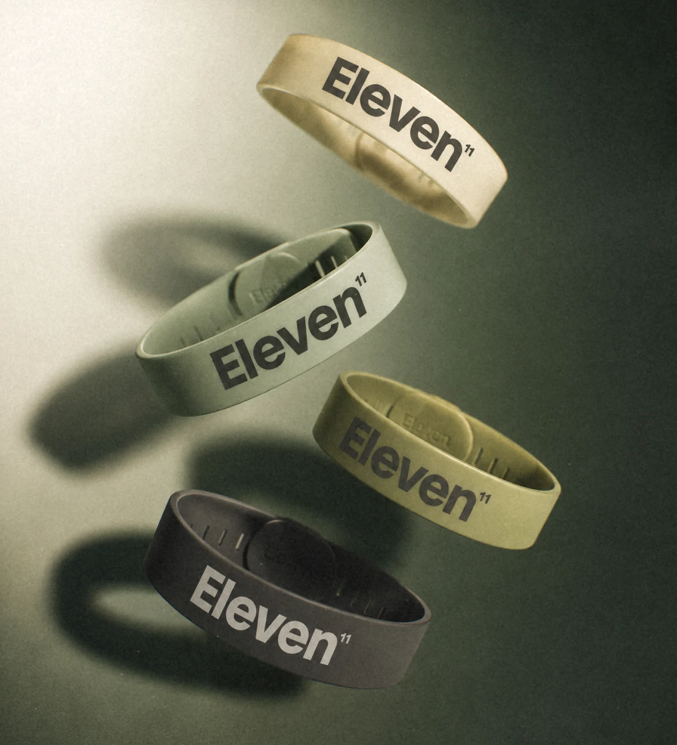 Four silicone wristbands in beige, gray, olive, and black with Eleven 11 text floating on a dark background.