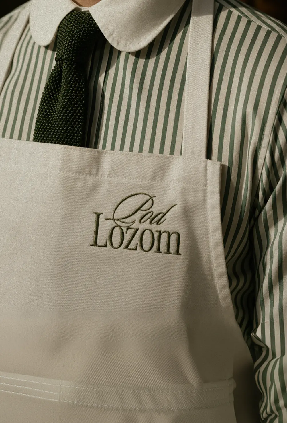 Close-up of a person wearing a green striped shirt, dark green knit tie, and beige apron with "Pod Lozom" text.