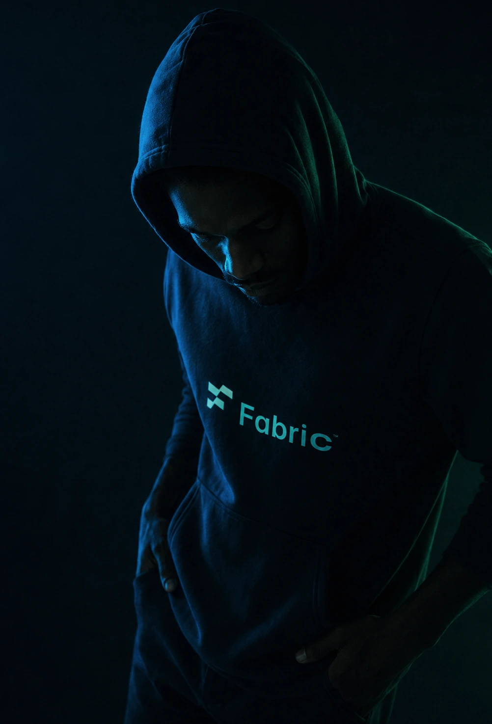 Man in dark hoodie with Fabric logo looking down in low light against dark background.