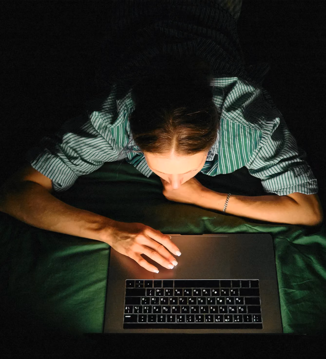 Overhead shot of a person typing on a laptop at night, wearing a green striped shirt.