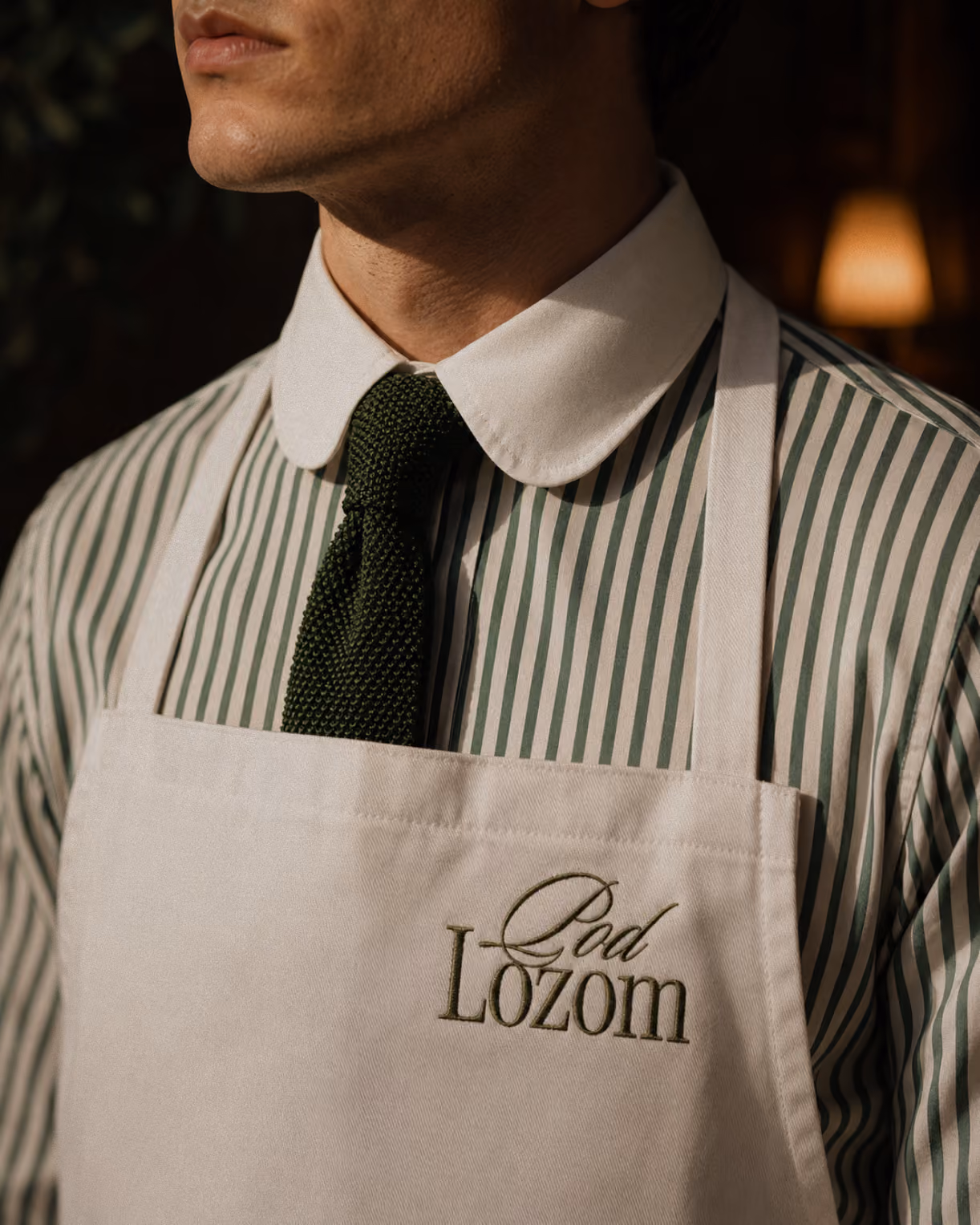 Man wearing a "Pod Lozom" branded apron over a striped shirt and green tie.