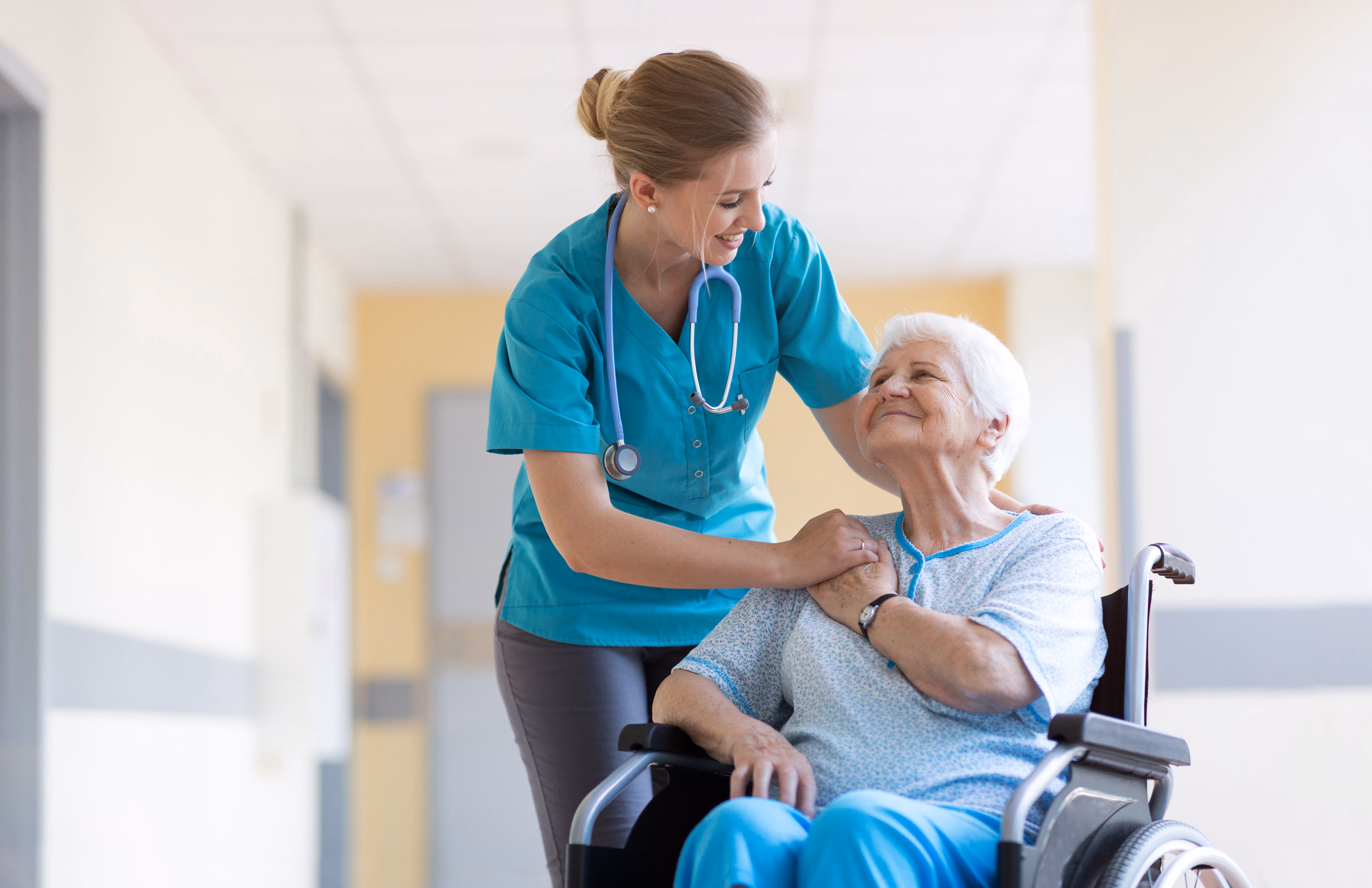 Healthcare professional assisting an older woman seated in a wheelchair in a care facility.