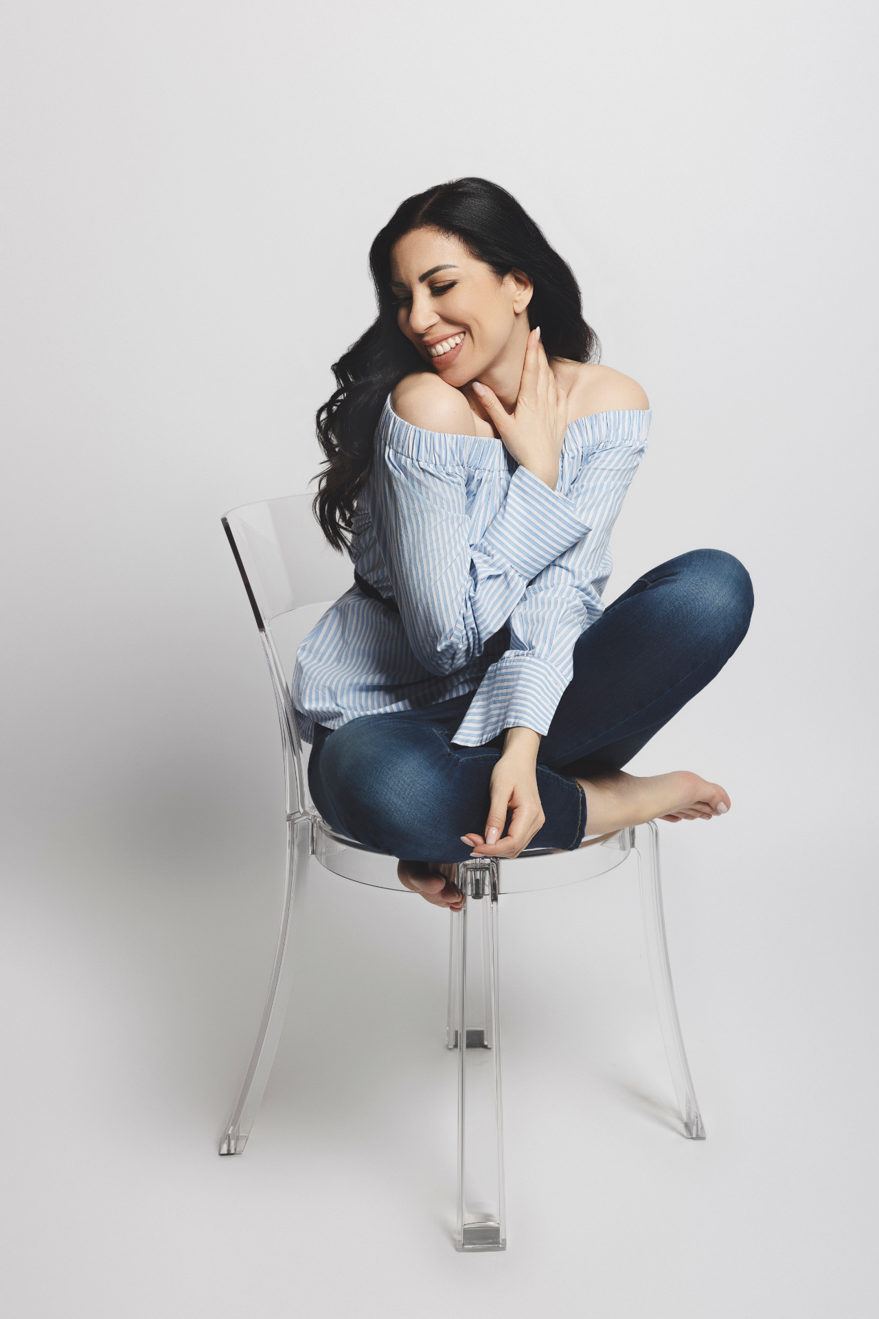 Rosy, cantante lirica, seduta a gambe incrociate su sedia trasparente in camicia a righe off-shoulder e jeans, ritratto casual per book di Claudia Spaziani a Castel Guelfo, Bologna.