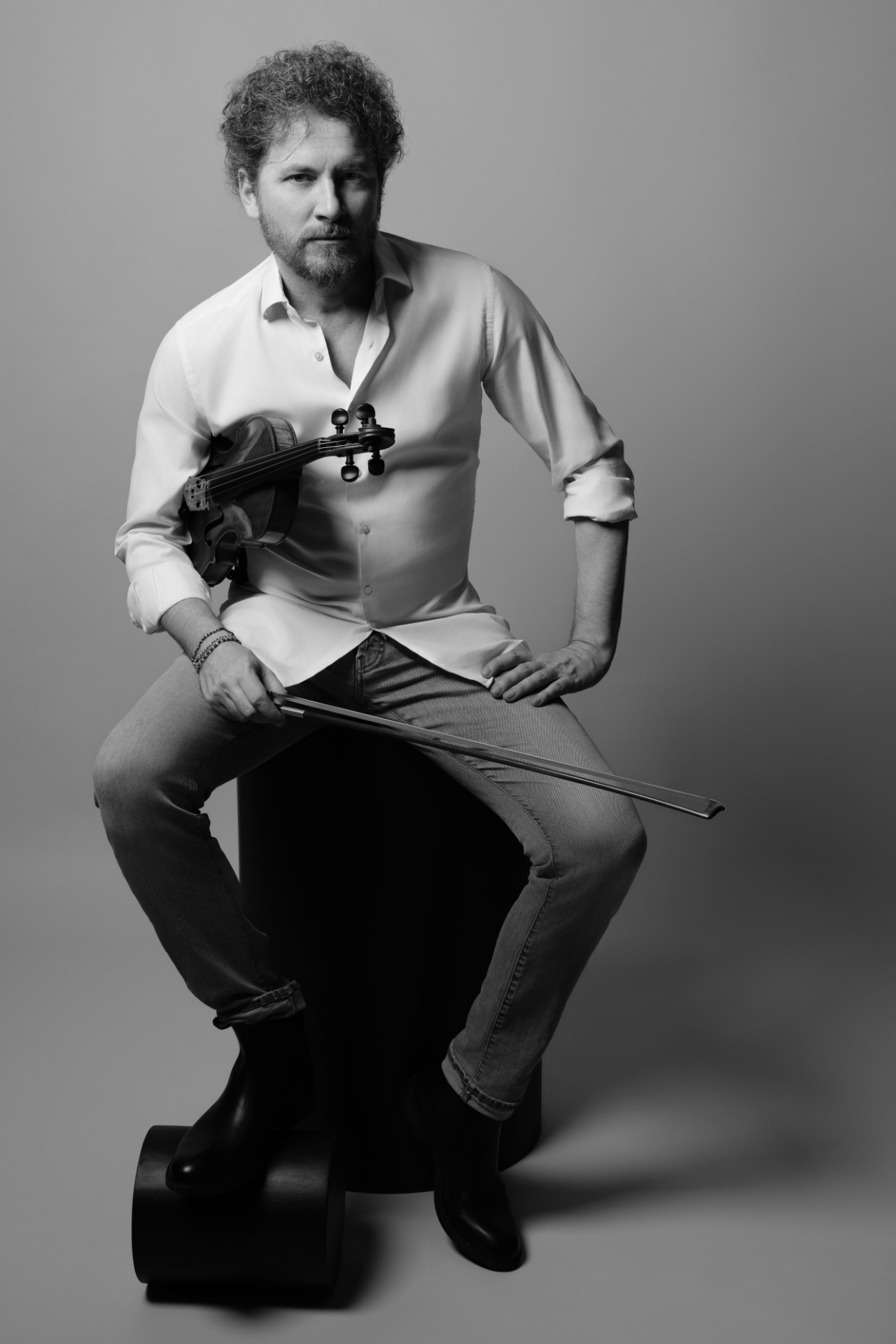 Violinista in bianco e nero seduto con violino e archetto in mano in camicia bianca e jeans, ritratto casual per book professionale di Claudia Spaziani a Castel Guelfo, Bologna.
