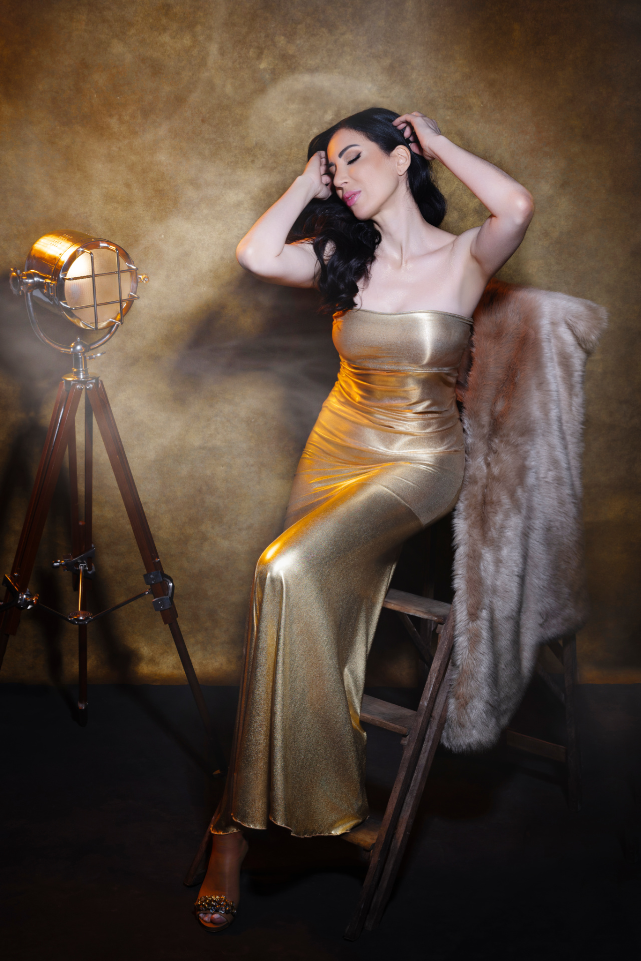 Rosy, cantante lirica, in abito dorato con pelliccia su scala in legno accanto a faro vintage, ritratto glamour per book professionale di Claudia Spaziani a Bologna.
