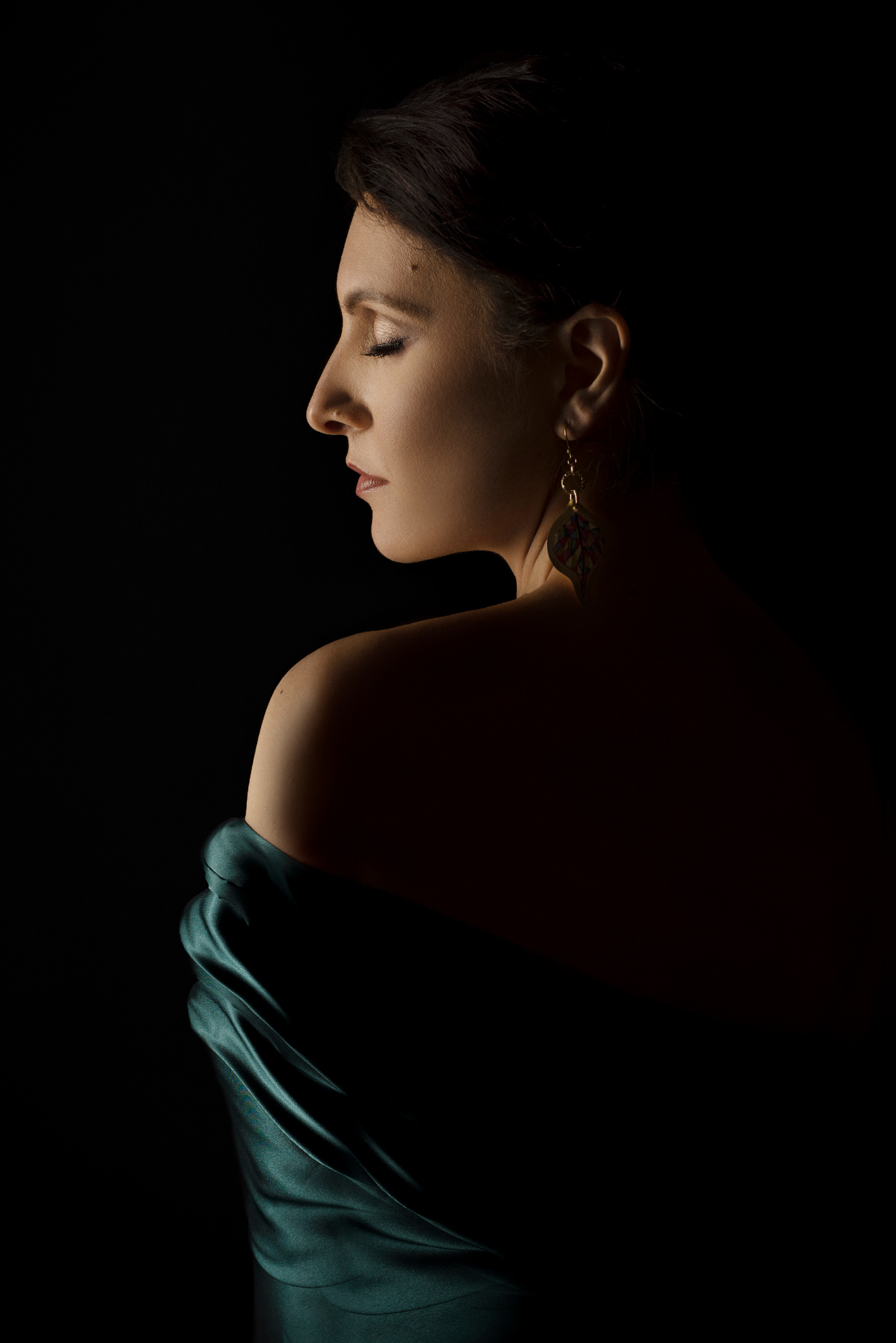 Luisa, soprano, di profilo con occhi chiusi e abito verde smeraldo off-shoulder, luce scultorea su sfondo nero, ritratto fine art di Claudia Spaziani a Castel Guelfo, Bologna.