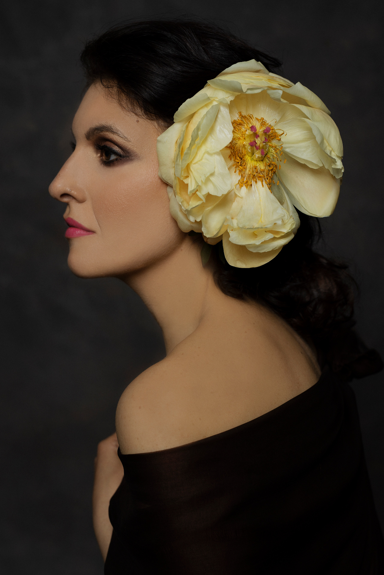 Luisa, cantante lirica, di profilo con grande peonia avorio tra i capelli scuri e abito off-shoulder nero, ritratto fine art per book professionale di Claudia Spaziani a Bologna.
