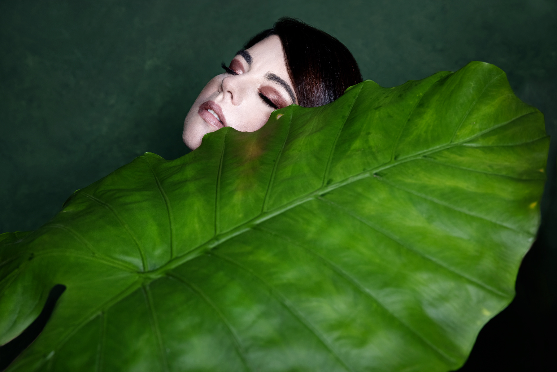 Ritratto fine art di Claudia Spaziani con elemento naturale; volto parzialmente nascosto da una grande foglia verde, atmosfera intensa e stilizzata.