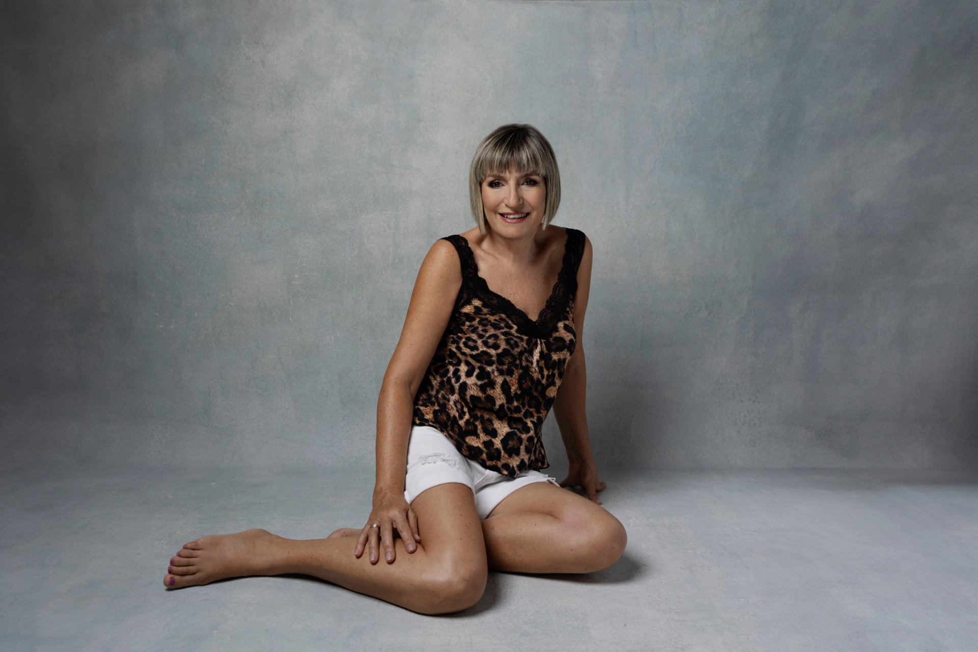 Ritratto glamour fine art di una donna seduta su sfondo grigio con top leopardato e shorts bianchi. Posa rilassata e composizione professionale. Fotografia realizzata da Claudia Spaziani nel suo studio fotografico a Bologna.