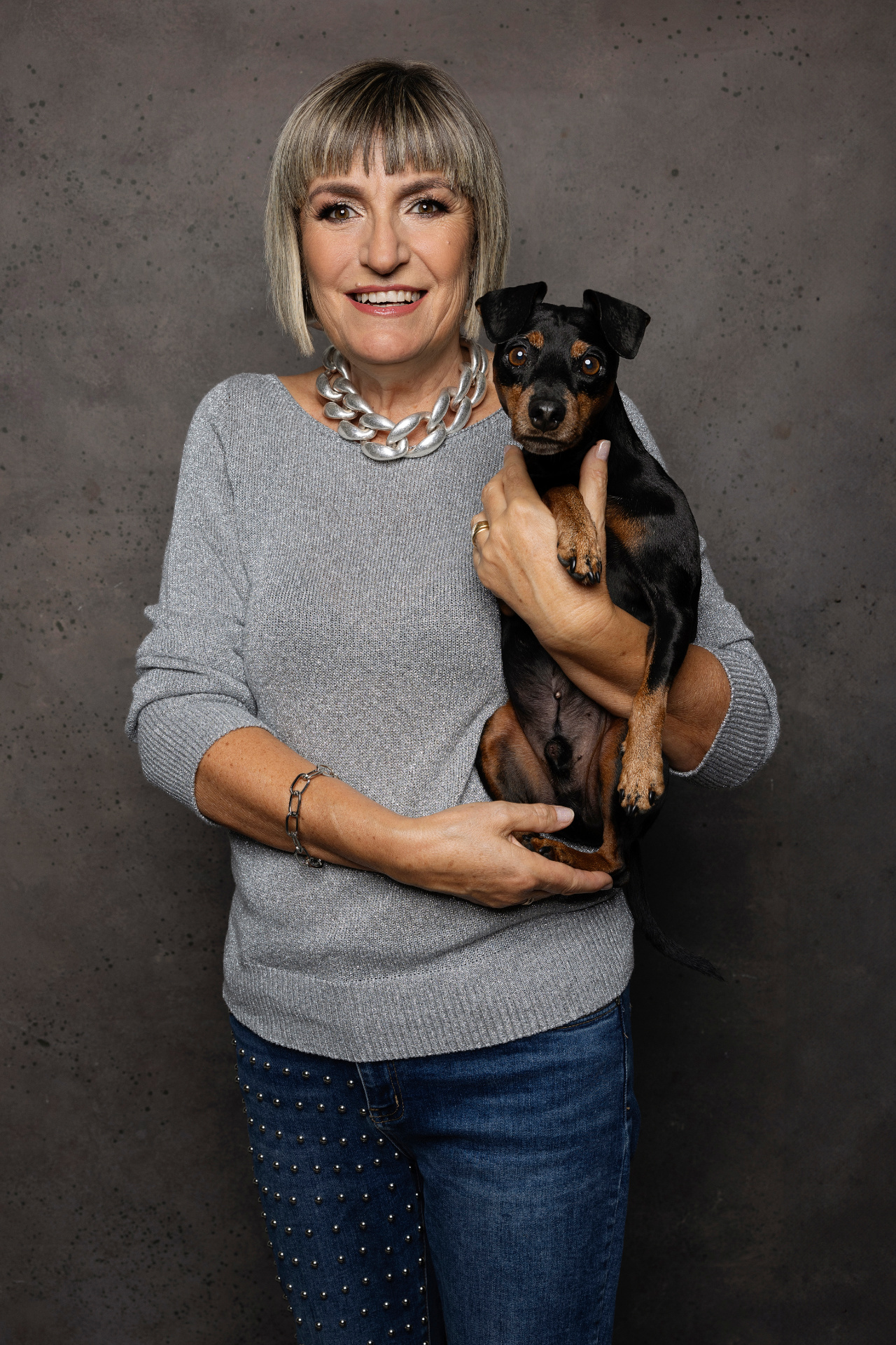 Donna con maglione grigio e collana argento tiene in braccio Miniature Pinscher nero focato su sfondo scuro. Ritratto fine art realizzato da Claudia Spaziani nel suo studio fotografico a Bologna.