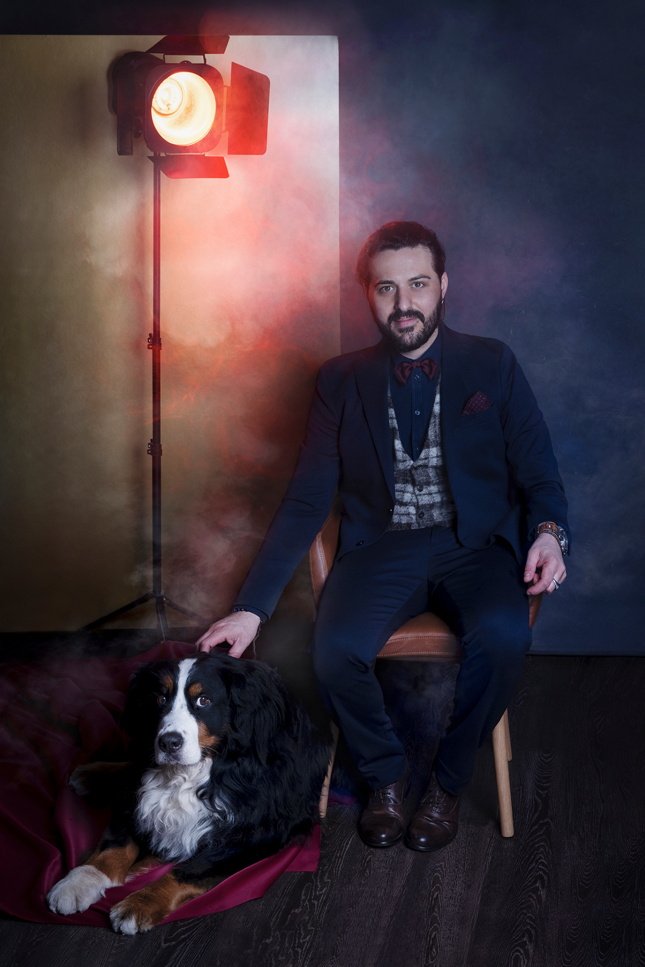 Uomo in abito formale seduto su sedia in legno accanto a Bovaro del Bernese sdraiato su tessuto bordeaux, con luce rossa e atmosfera teatrale. Ritratto fine art realizzato da Claudia Spaziani nel suo studio a Bologna.