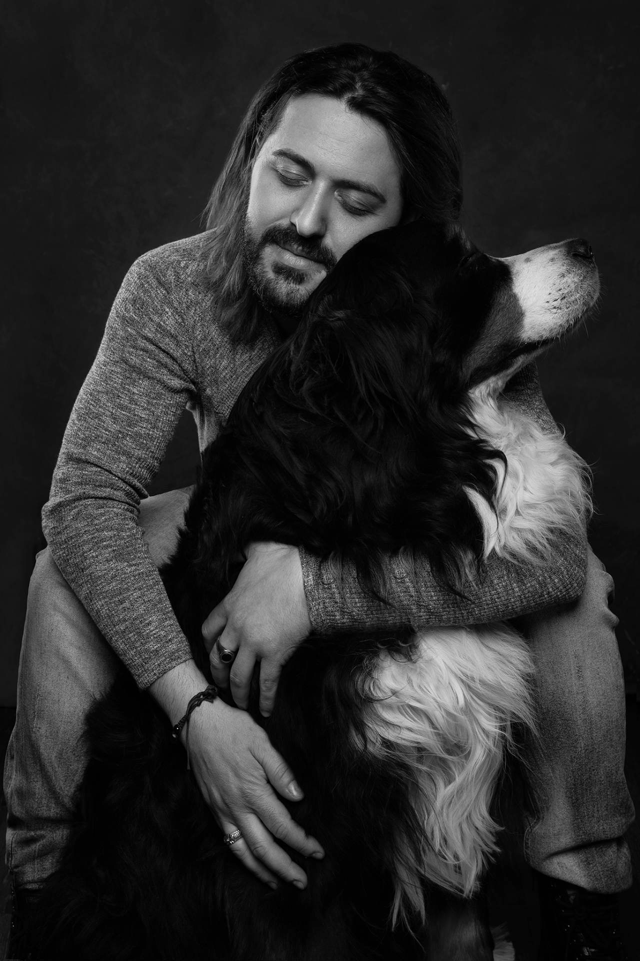 Fotografia in bianco e nero di uomo con barba e capelli lunghi che abbraccia un Bovaro del Bernese, esprimendo profonda tenerezza. Ritratto fine art di Claudia Spaziani, Bologna.