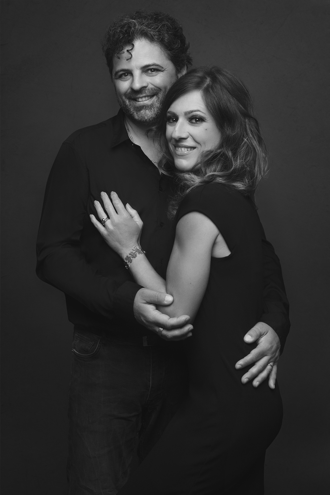 Fotografia fine art in bianco e nero di coppia in posa ravvicinata, scattata da Claudia Spaziani a Bologna. Lei in abito scuro senza maniche, lui in camicia nera e jeans, entrambi sorridenti su sfondo neutro.
