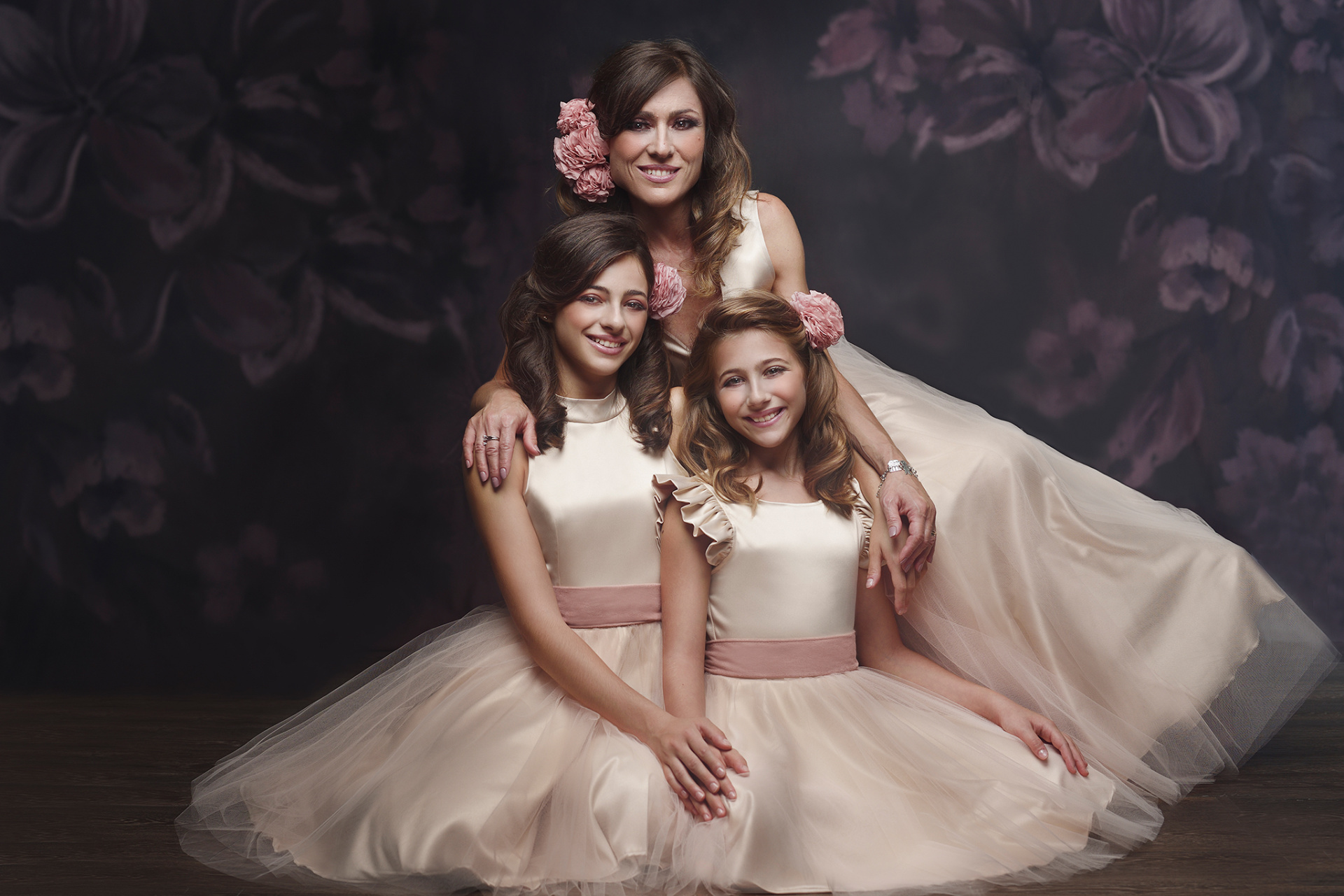 Fotografia fine art di madre e due figlie in abiti color crema con dettagli rosa, scattata da Claudia Spaziani a Bologna. Posa abbracciata su sfondo floreale scuro, con accessori coordinati nei capelli.