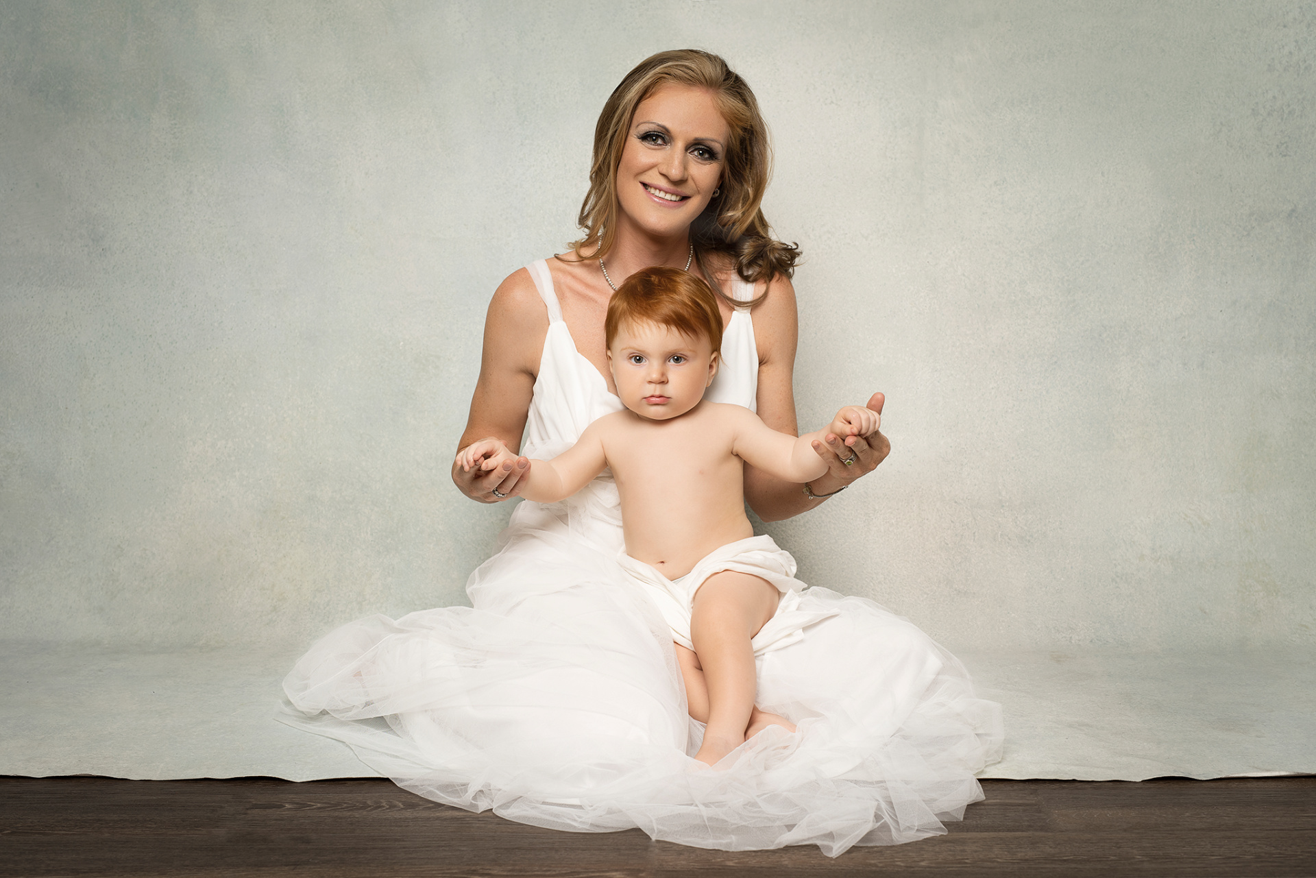 Fotografia fine art di madre e figlio seduti su pavimento, scattata da Claudia Spaziani nel suo studio a Bologna. Lei in abito bianco vaporoso, lui in pannolino bianco, in posa affettuosa su sfondo neutro.