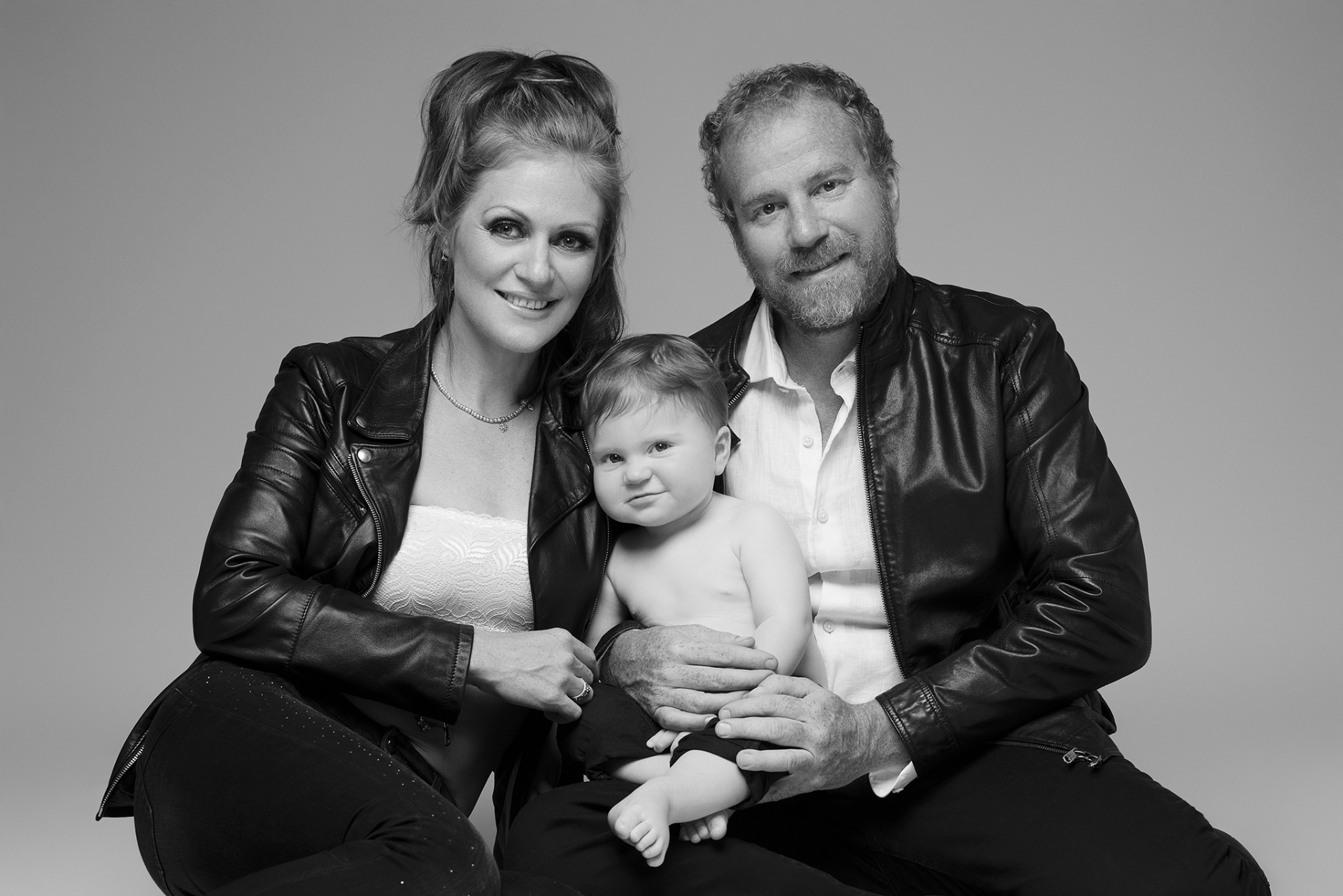Fotografia fine art in bianco e nero di madre, padre e figlio, scattata da Claudia Spaziani nel suo studio a Bologna. Genitori in giacche di pelle nere, bambino al centro in posa affettuosa su sfondo neutro.