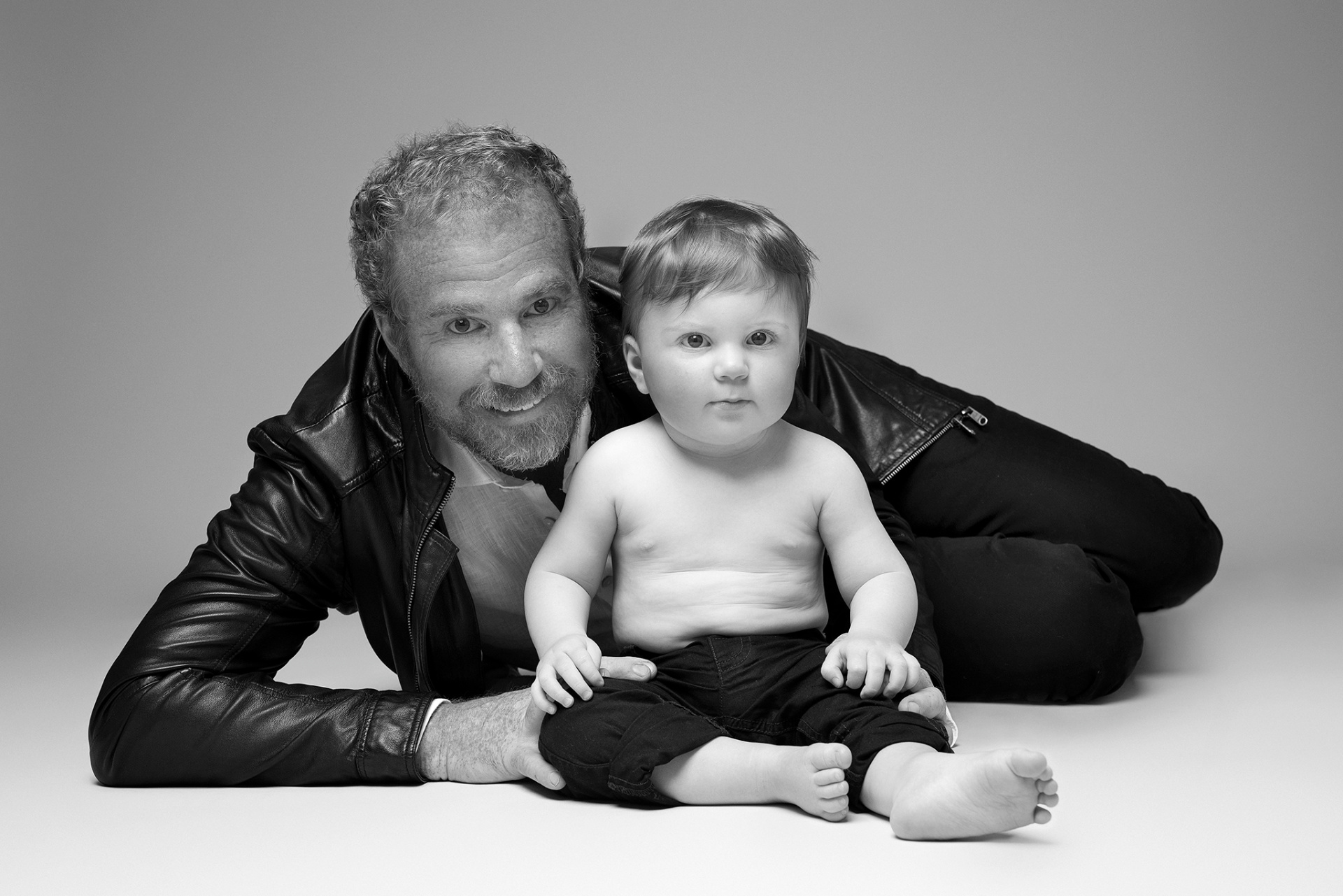 Fotografia fine art in bianco e nero di padre e figlio, scattata da Claudia Spaziani nel suo studio a Bologna. Padre sdraiato con giacca di pelle e sorriso, figlio seduto davanti in pantaloni scuri, su sfondo neutro.