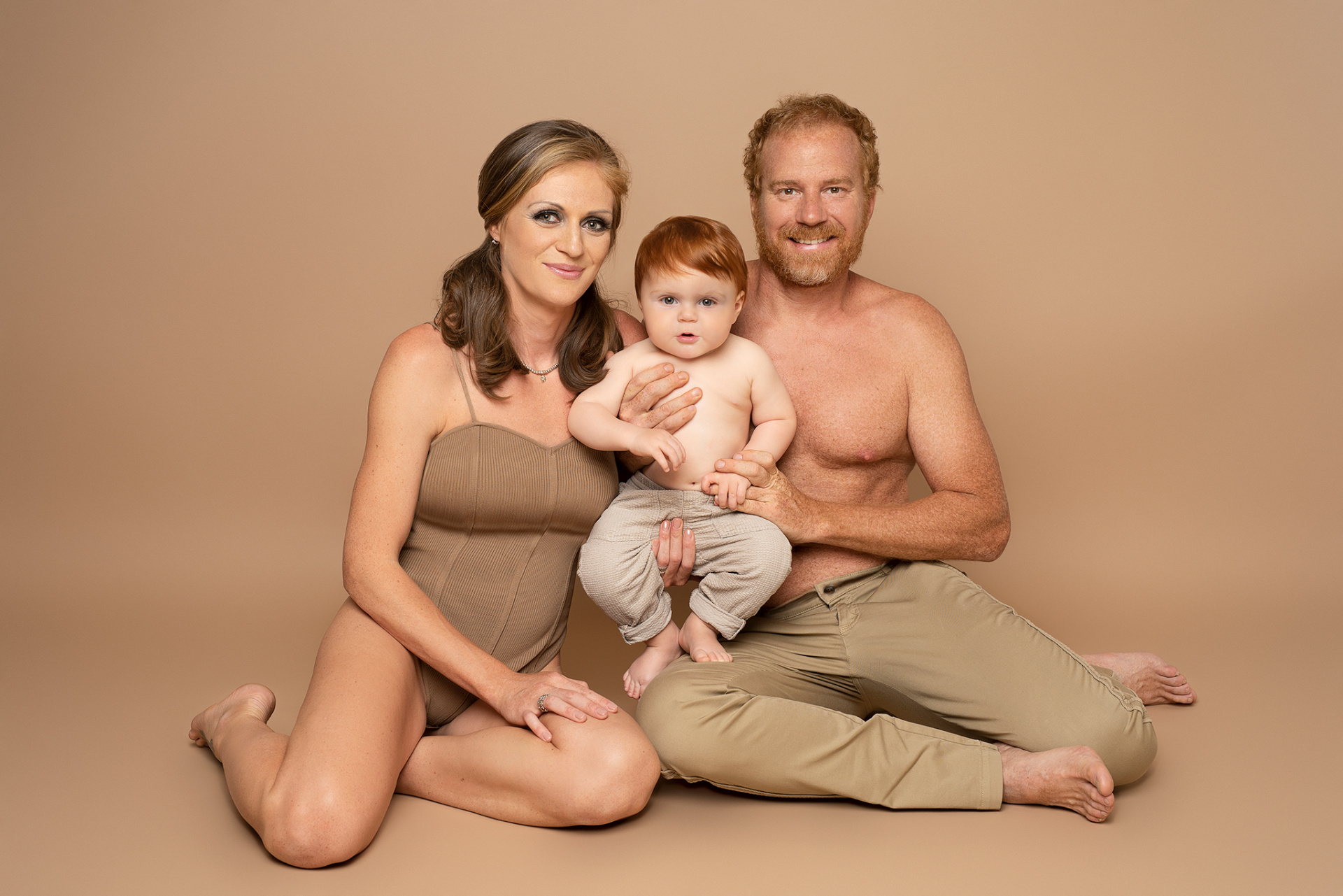 Ritratto artistico di madre, padre e figlio in abiti coordinati beige, realizzato da Claudia Spaziani a Bologna. Composizione calda e armoniosa su pavimento chiaro, con il bambino al centro.