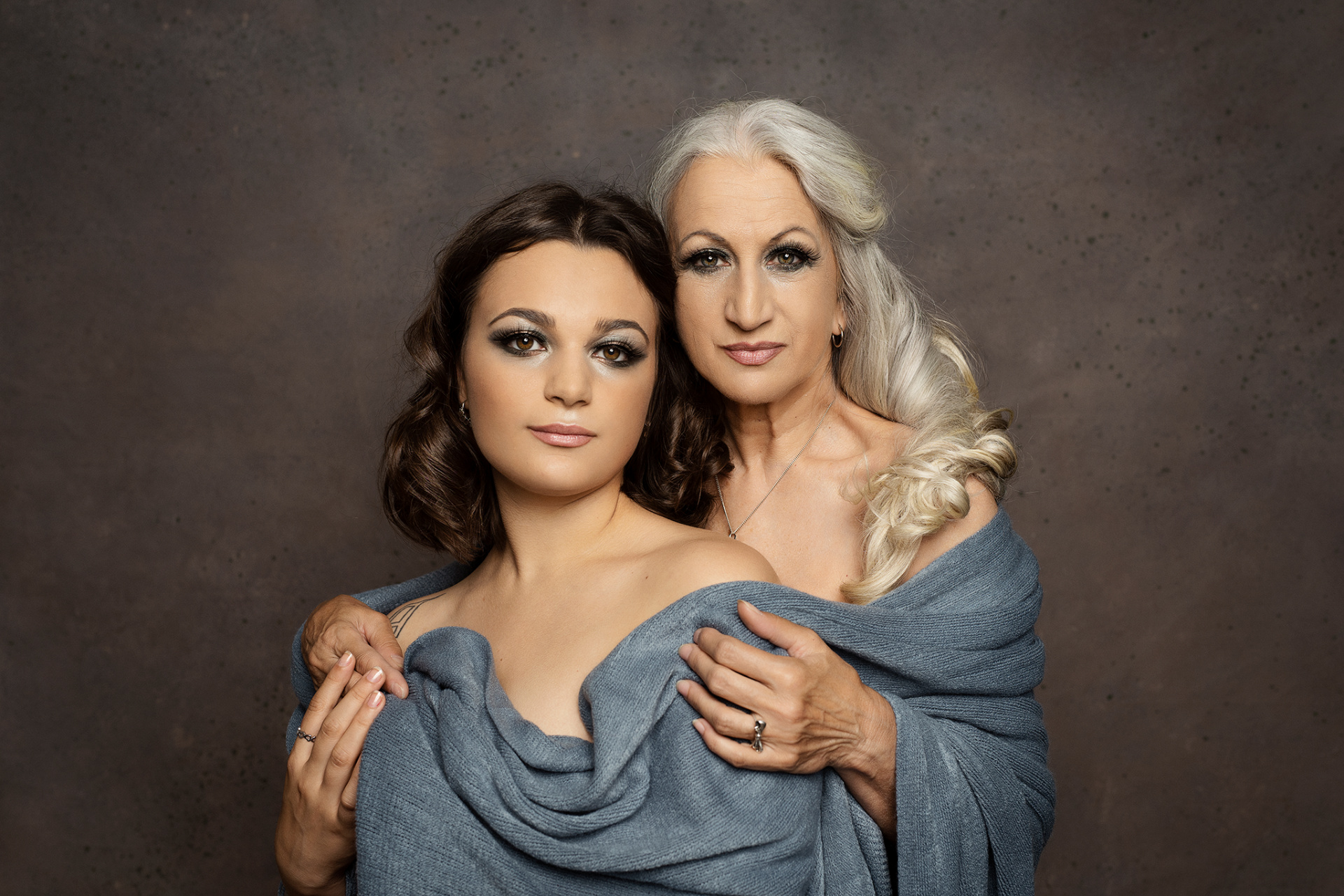 Fotografia fine art di madre e figlia avvolte in tessuto blu-grigio, scattata da Claudia Spaziani nel suo studio a Bologna. Posa ravvicinata su sfondo neutro, con espressione intensa e legame affettuoso.