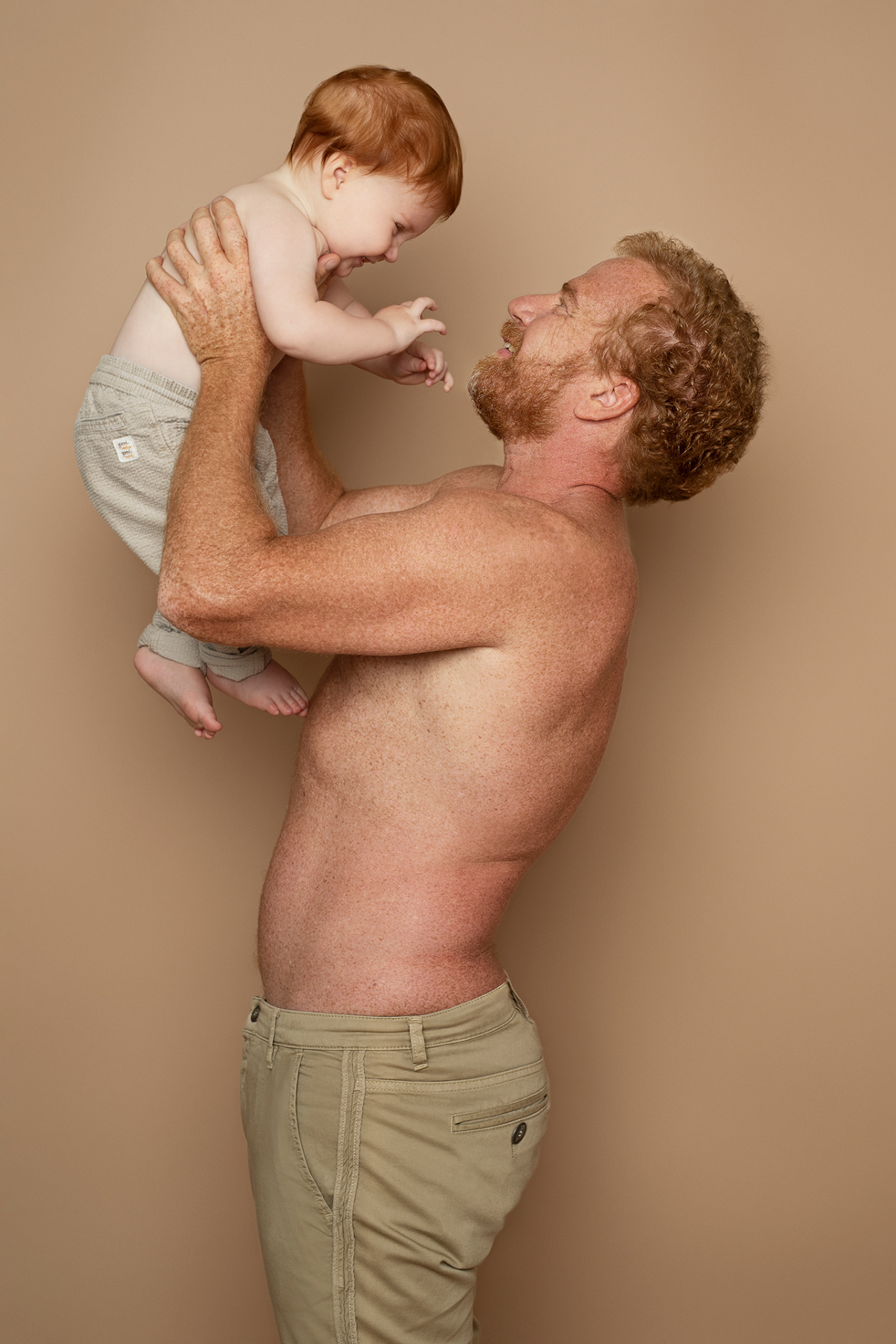 Fotografia fine art di padre e figlio con capelli rossi, scattata da Claudia Spaziani nel suo studio a Bologna. Posa giocosa su sfondo beige, con il bambino sollevato in aria e mani tese.