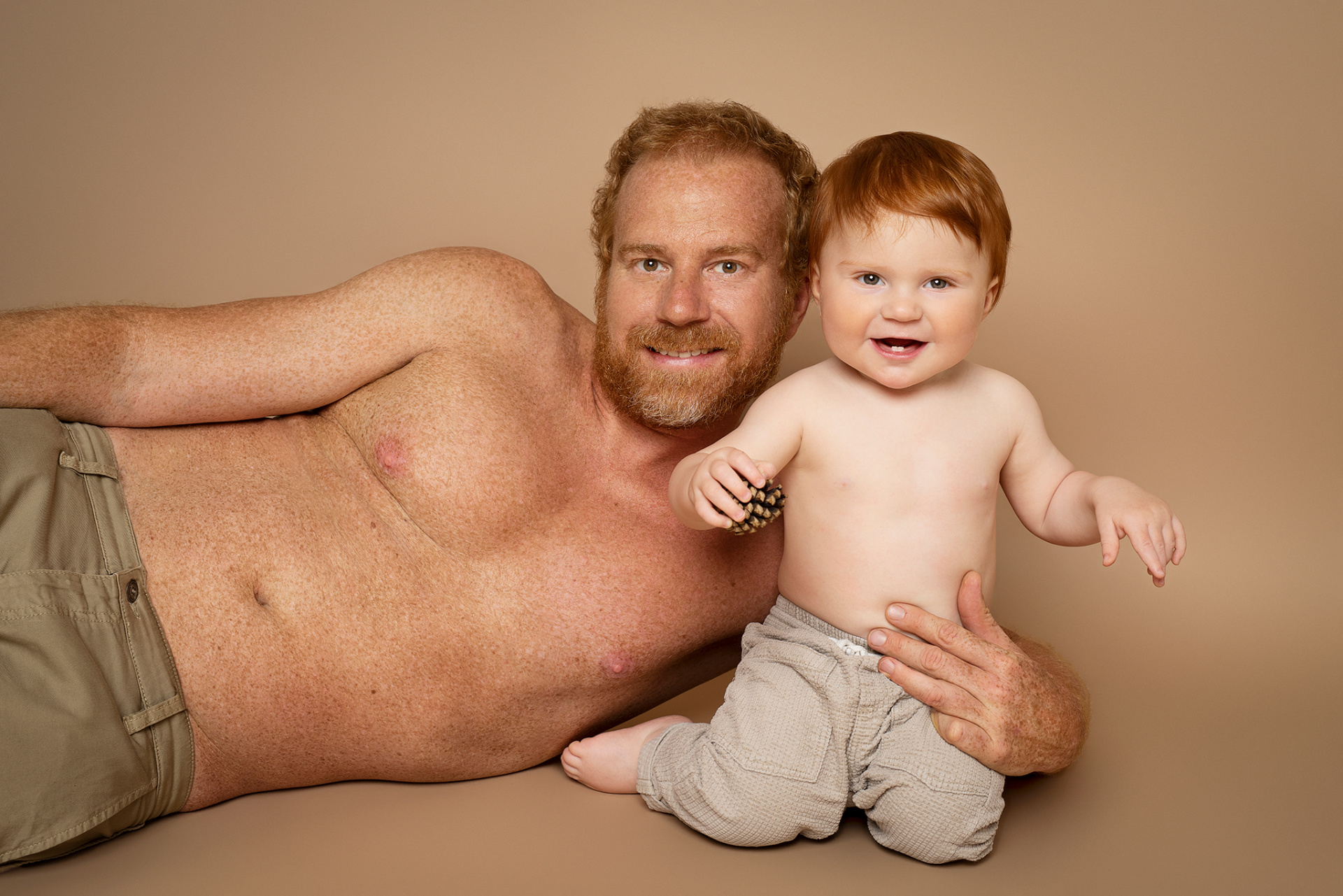Fotografia fine art di padre e figlio in posa affettuosa, scattata da Claudia Spaziani nel suo studio a Bologna. Entrambi a torso nudo con pantaloni beige, il bambino tiene una pigna.