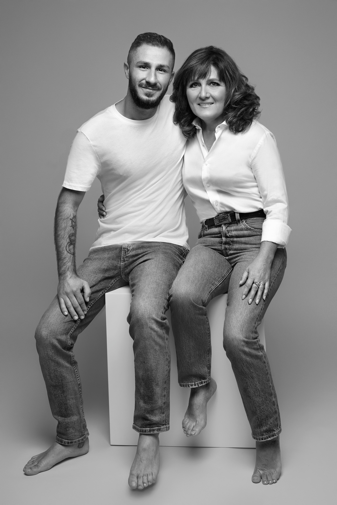 Fotografia fine art in bianco e nero di madre e figlio sedute su cubo bianco, scattata da Claudia Spaziani nel suo studio a Bologna. Entrambi in jeans e camicie bianche, a piedi nudi, in posa ravvicinata.