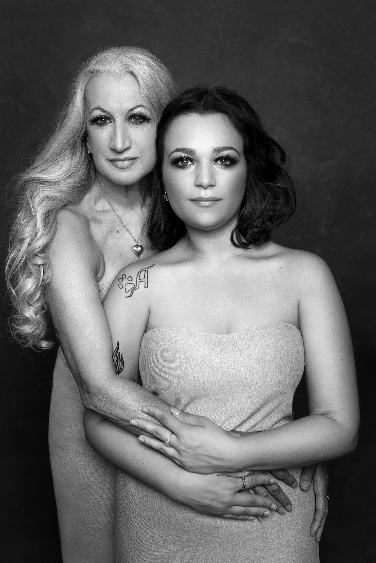 Ritratto artistico in bianco e nero di madre e figlia in abbraccio, realizzato da Claudia Spaziani a Bologna. Espressione serena, tatuaggi visibili e collana con ciondolo a cuore su sfondo scuro.