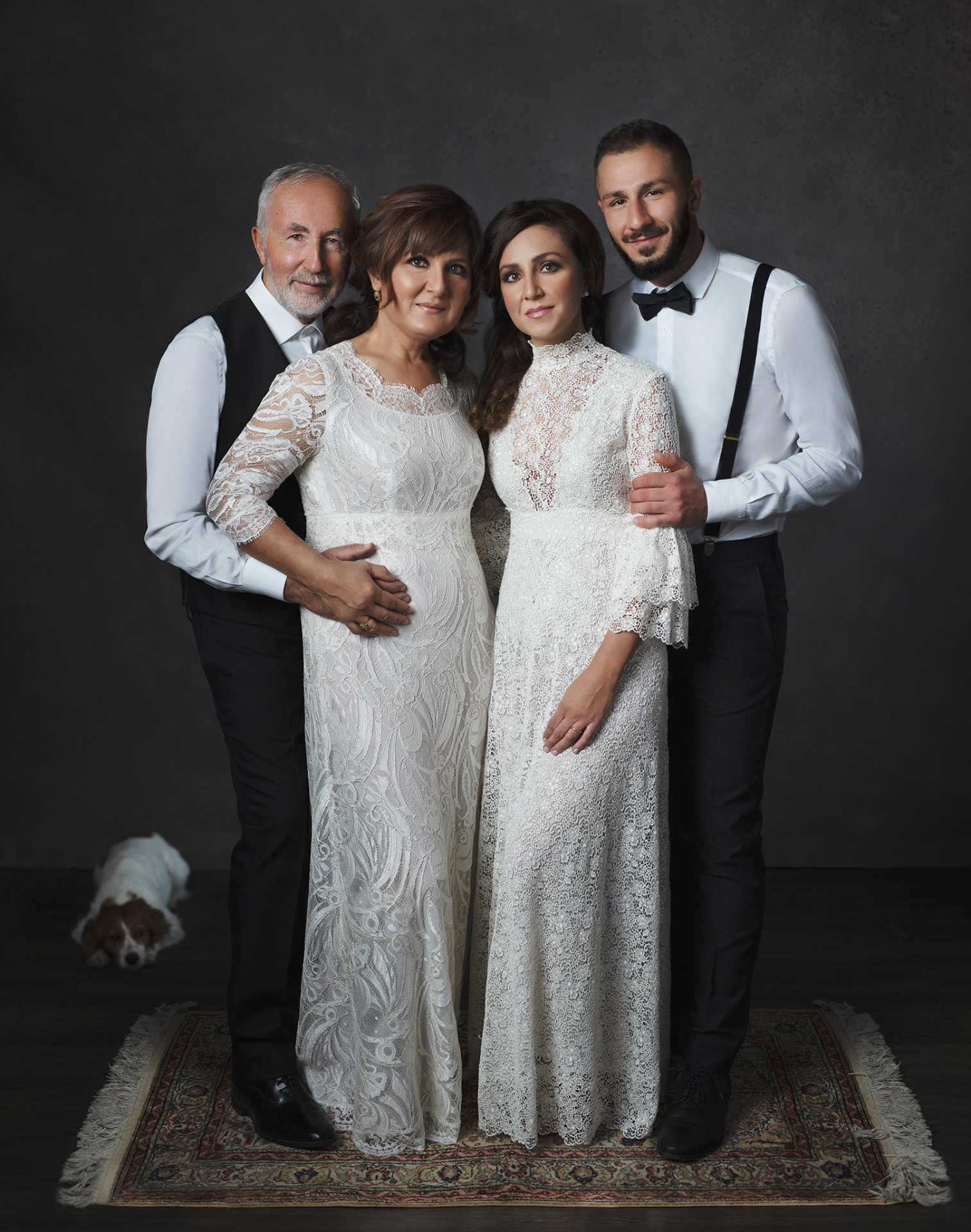 Ritratto artistico di famiglia con quattro adulti e un cane, realizzato da Claudia Spaziani a Bologna. Donne in abiti bianchi in pizzo, uomini in camicia bianca con bretelle nere e papillon, su tappeto decorativo.