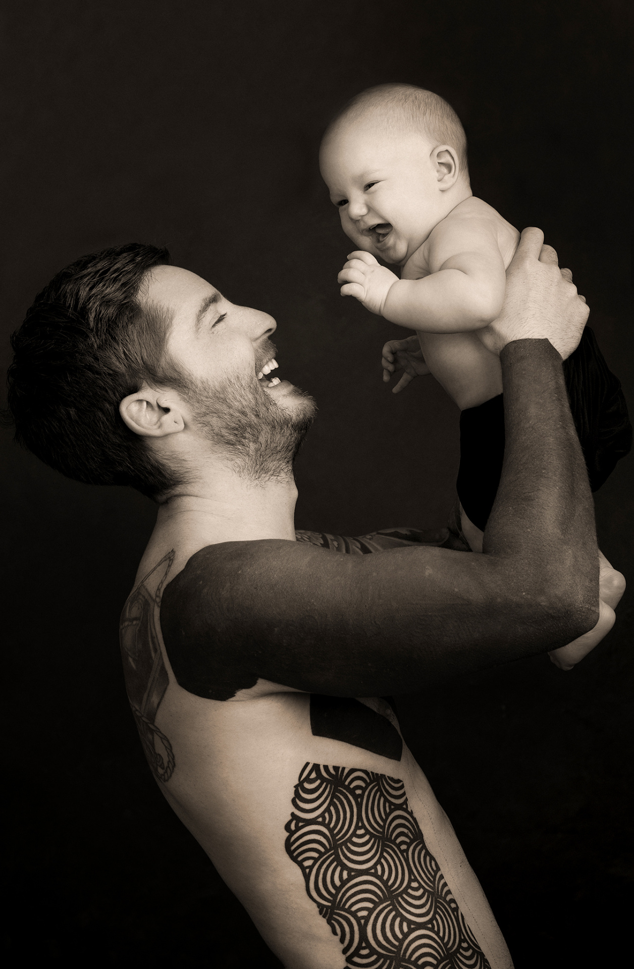 Ritratto in seppia: padre a torso nudo con tatuaggi solleva fra le braccia il figlio neonato che ride. Fotografia di famiglia di Claudia Spaziani, Bologna.