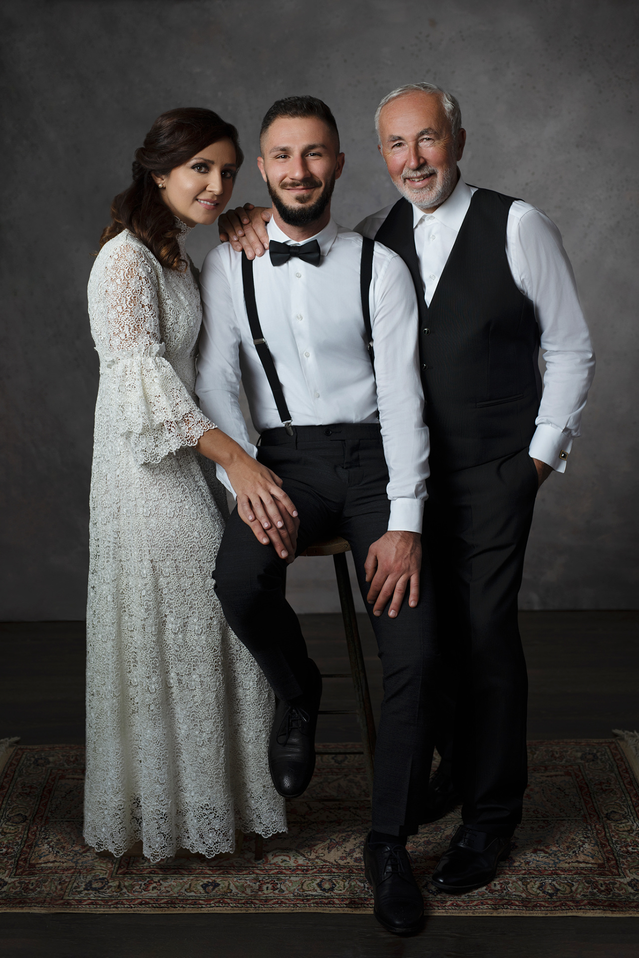 Fotografia artistica di padre con i suoi figli in abiti da cerimonia, realizzata da Claudia Spaziani a Bologna. Donna in abito bianco in pizzo, uomo in camicia con bretelle e papillon, terzo in gilet nero, su sfondo neutro.