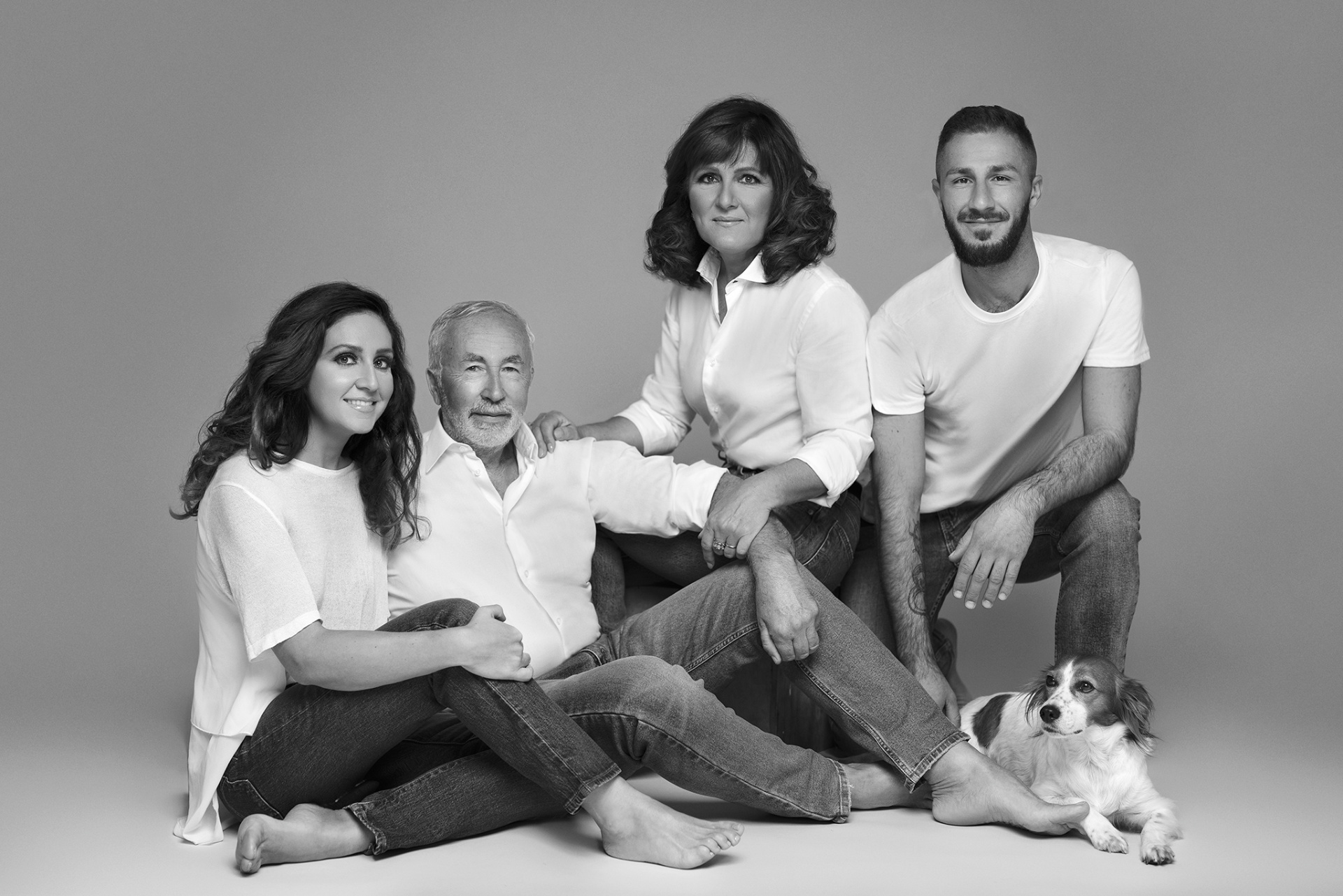 Ritratto in studio in bianco e nero di famiglia con cane, realizzato da Claudia Spaziani a Bologna. Quattro persone in jeans e maglietta bianca, sedute e inginocchiate su sfondo neutro, con cane disteso in primo piano.