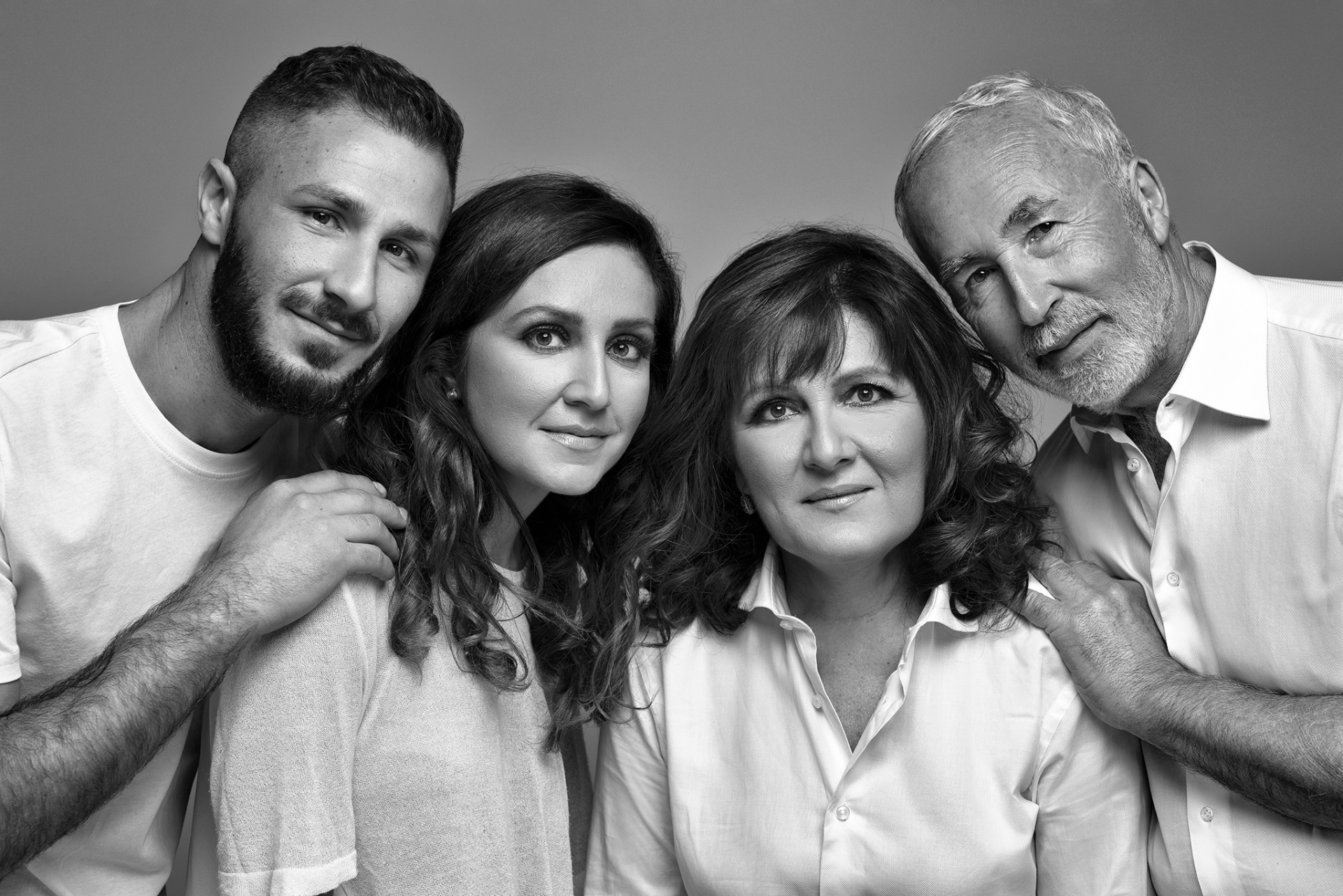 Ritratto fine art in bianco e nero di famiglia in abiti chiari, scattato da Claudia Spaziani nel suo studio a Bologna. Posa ravvicinata e gesti affettuosi su sfondo neutro.a