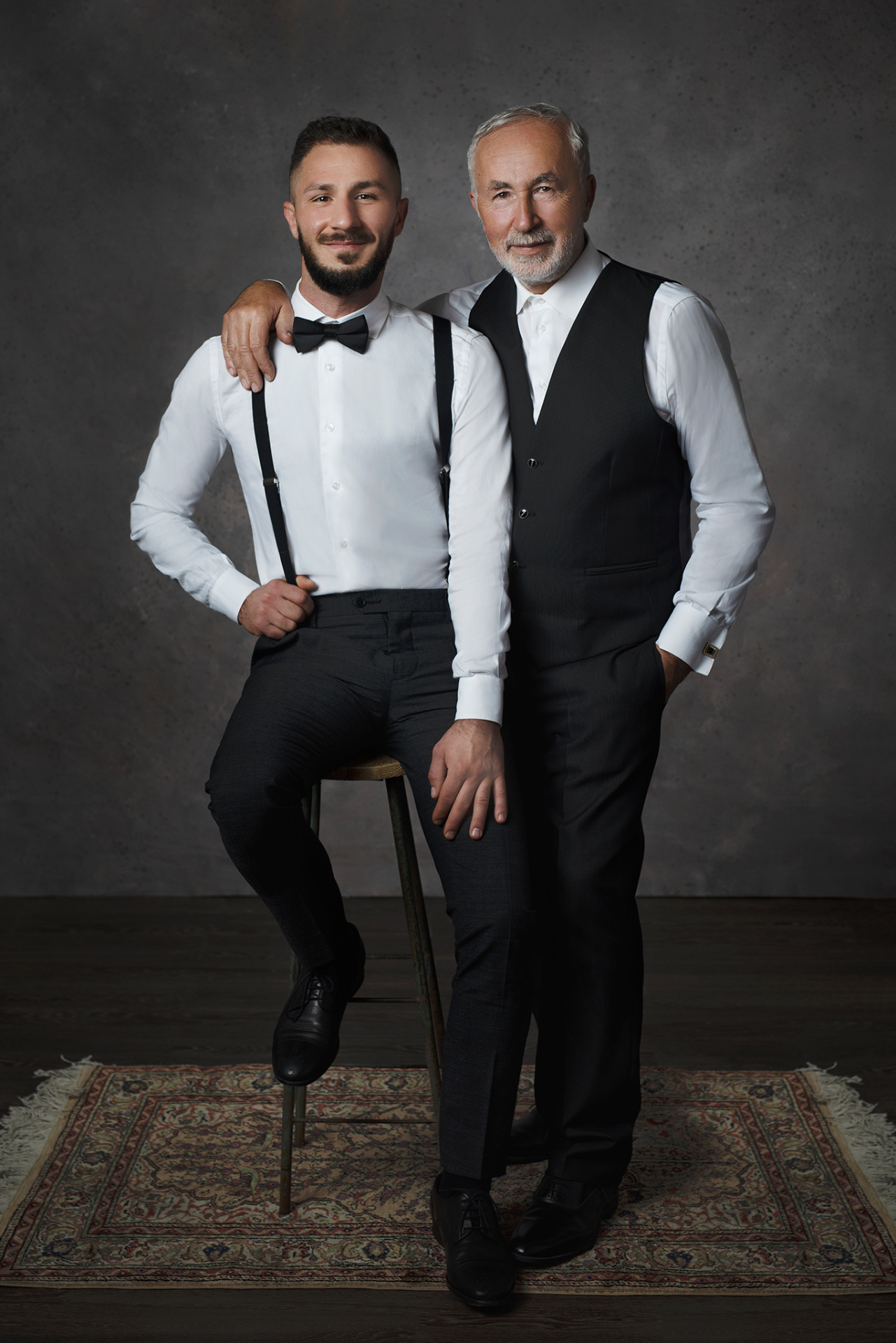 Fotografia fine art di padre e figlio in abiti eleganti, realizzata da Claudia Spaziani nel suo studio fotografico a Bologna. Uno seduto con bretelle e papillon, l’altro in piedi con gilet nero, su tappeto decorativo.