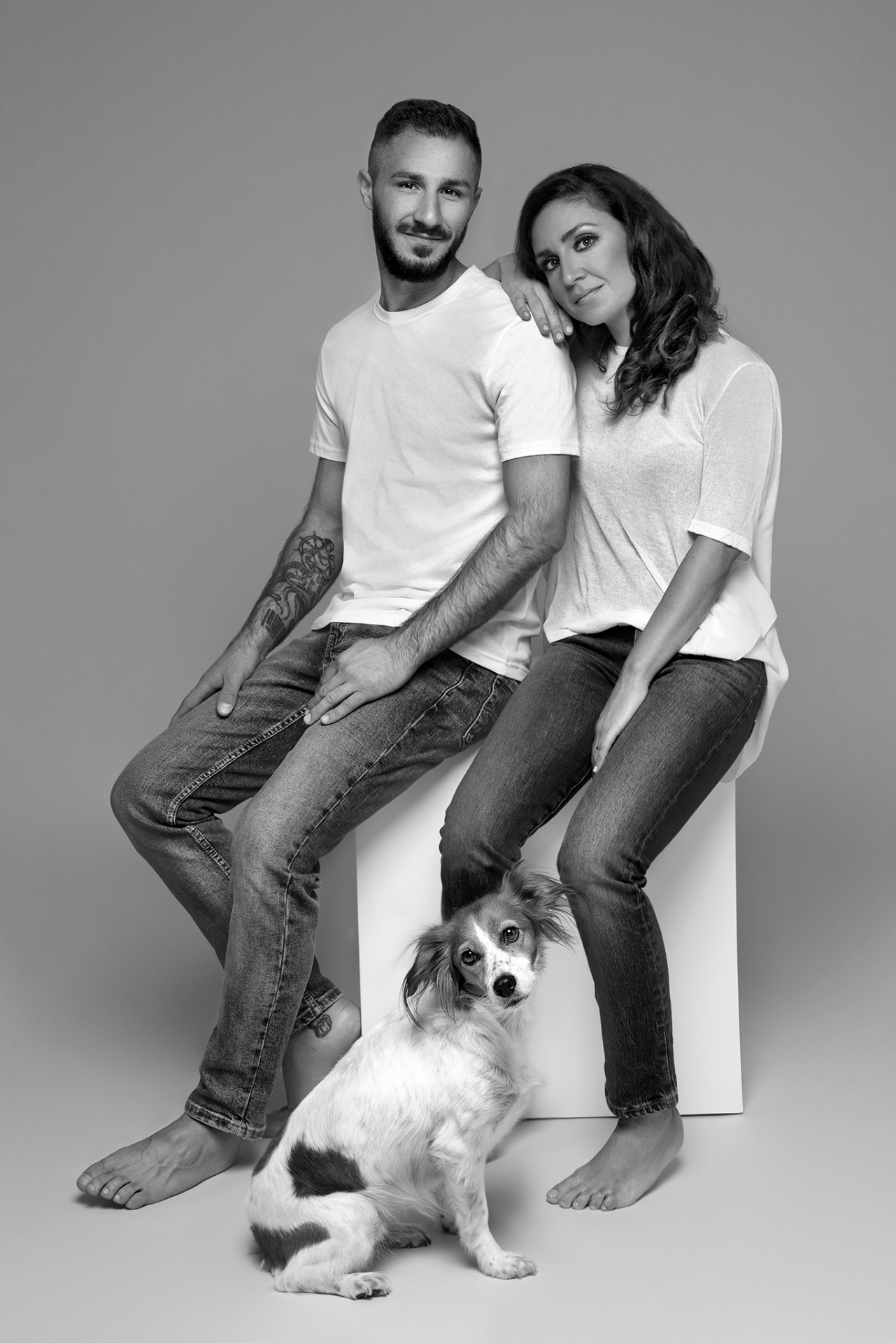 Ritratto fine art in bianco e nero di due fratelli con cane, scattato da Claudia Spaziani nel suo studio a Bologna. Entrambi in jeans e maglietta bianca, seduti su cubo bianco, con cane in primo piano.