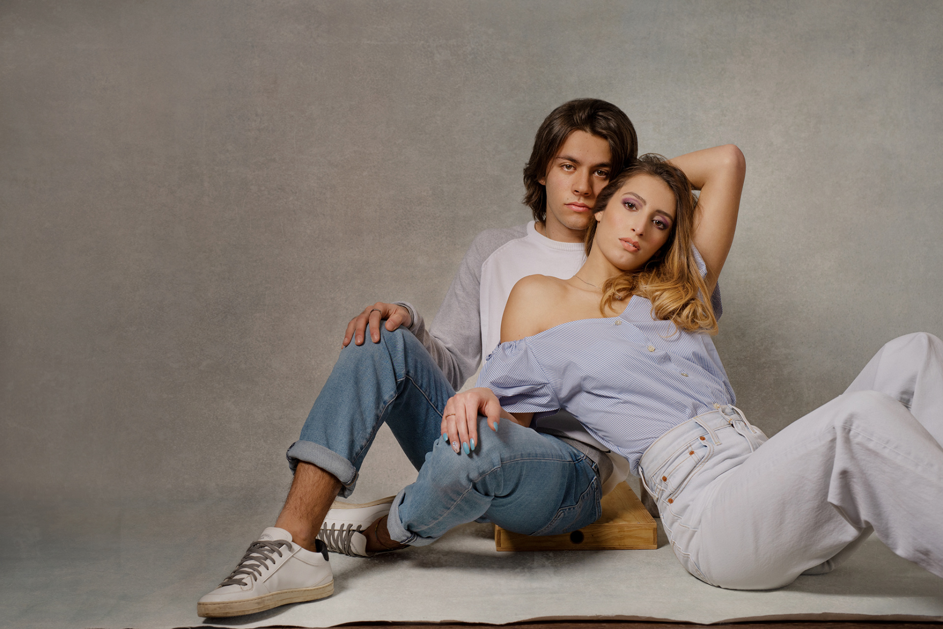 Ritratto moderno di una coppia in abbigliamento casual, seduta insieme in studio con sfondo grigio. Fotografia realizzata da Claudia Spaziani a Bologna, che evidenzia il legame affettivo attraverso una posa naturale e armoniosa.