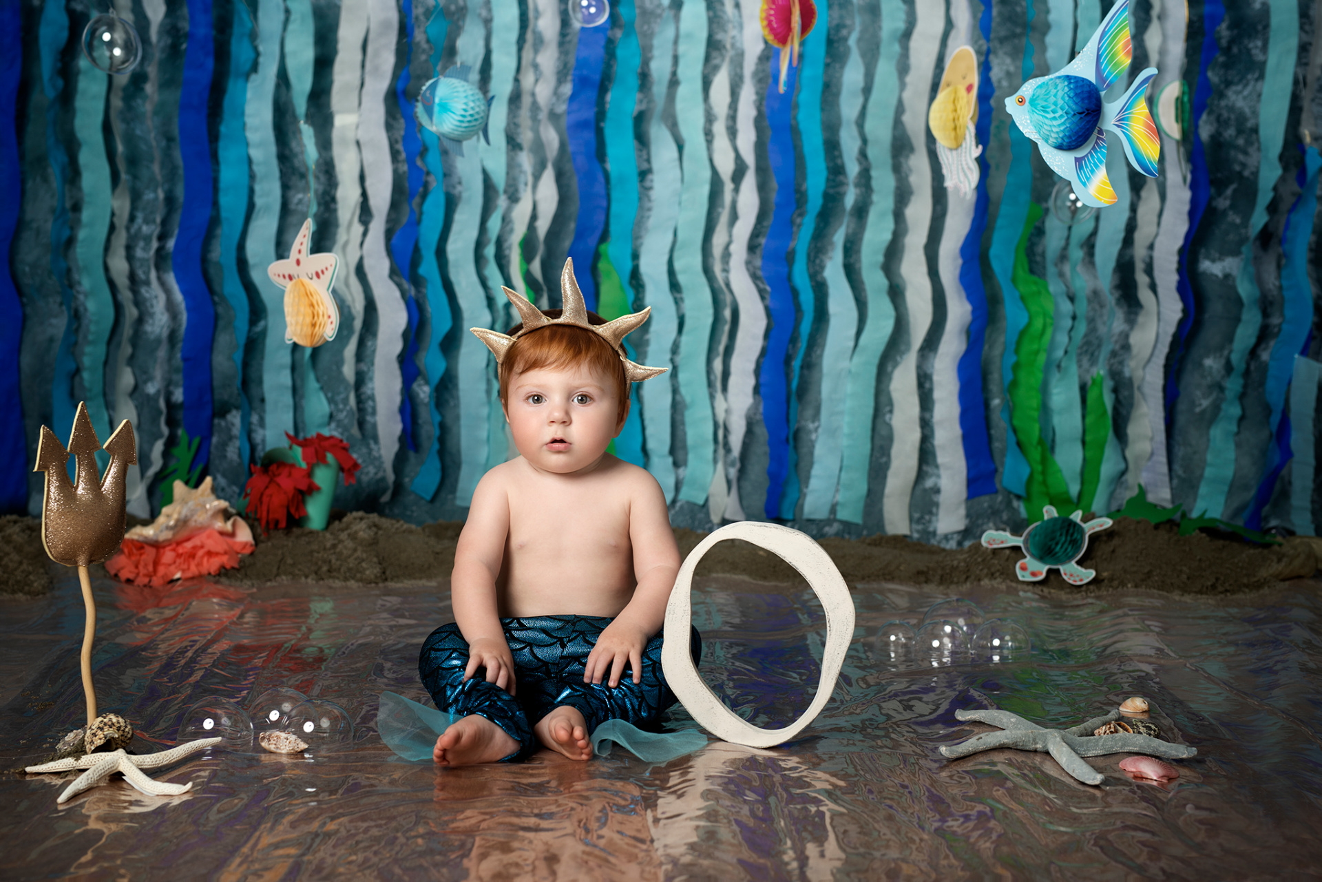 Bambino con pantaloni a squame blu e corona dorata a forma di corallo, seduto su superficie riflettente. Sfondo marino con pesci, meduse e stelle marine. Ritratto creativo di primo compleanno ispirato alla Sirenetta, scattato da Claudia Spaziani a Bologna.