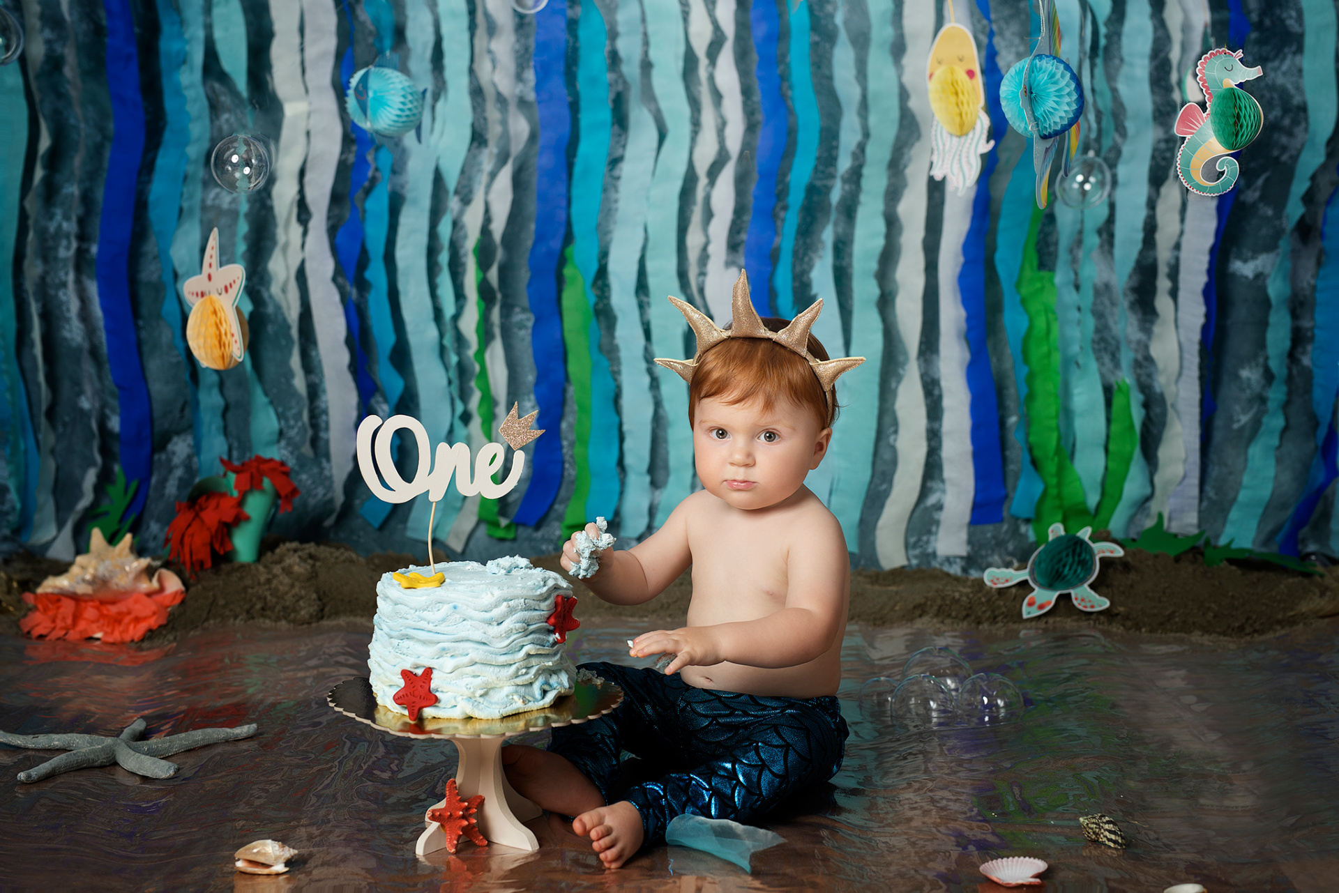 Bambino con pantaloni a squame blu e corona corallo, seduto davanti a torta azzurra con topper "One" e stelle marine. Sfondo marino con creature acquatiche e decorazioni subacquee. Ritratto di smash cake ispirato alla Sirenetta, realizzato da Claudia Spaziani a Bologna.