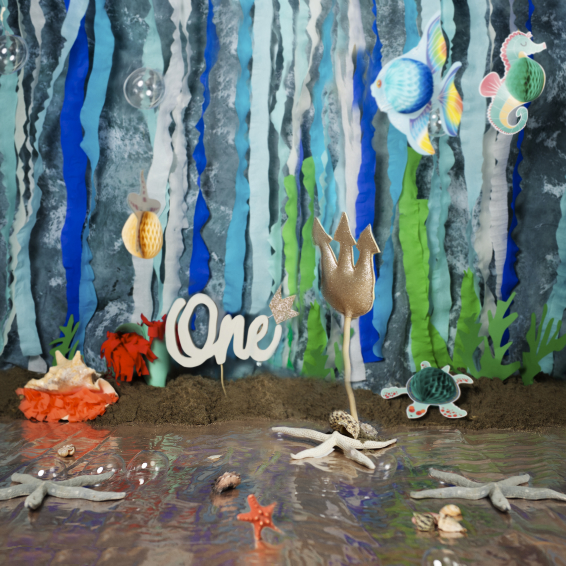 Allestimento fotografico a tema marino con decorazioni di pesci, cavallucci marini, granchio e alghe. Sfondo con nastri blu e verdi, tridente dorato e scritta "One". Ritratto di primo compleanno ispirato alla Sirenetta, realizzato da Claudia Spaziani nel suo studio fotografico a Bologna.