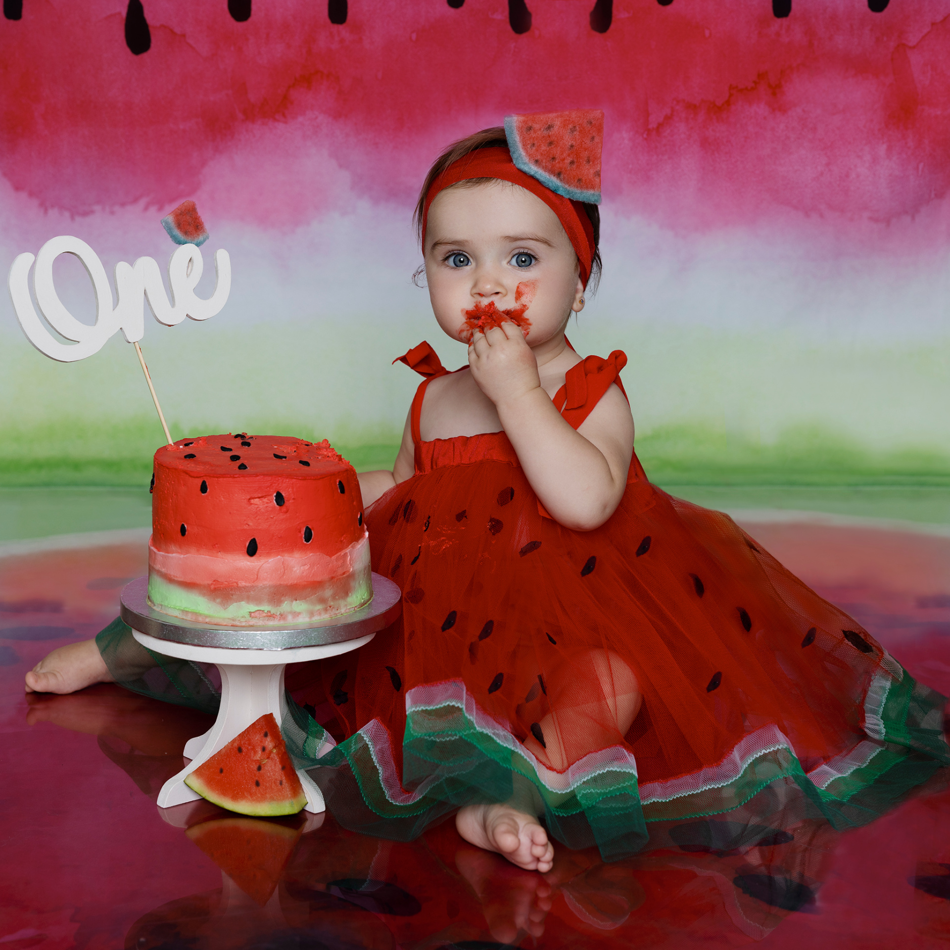 Bambina con vestito a tema anguria e fascia decorativa, seduta davanti a torta anguria con topper "One". Sfondo colorato con motivi di anguria. Foto di primo compleanno scattata da Claudia Spaziani nel suo studio fotografico a Bologna.