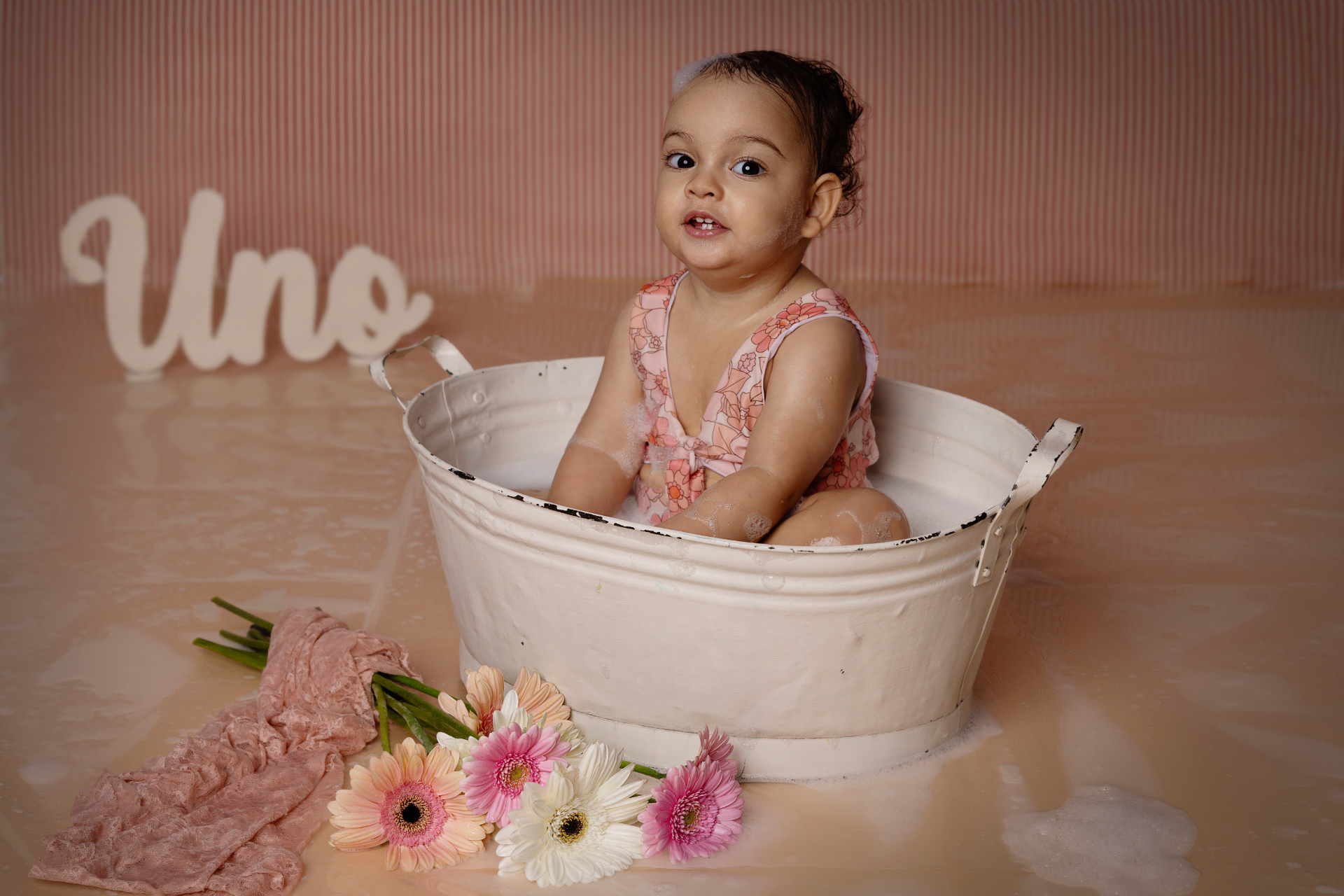 Bambina in vasca bianca con bolle di sapone e outfit rosa floreale, davanti a scritta "Uno" e fiori colorati. Ritratto di primo compleanno in stile bagno creativo, realizzato da Claudia Spaziani a Bologna.