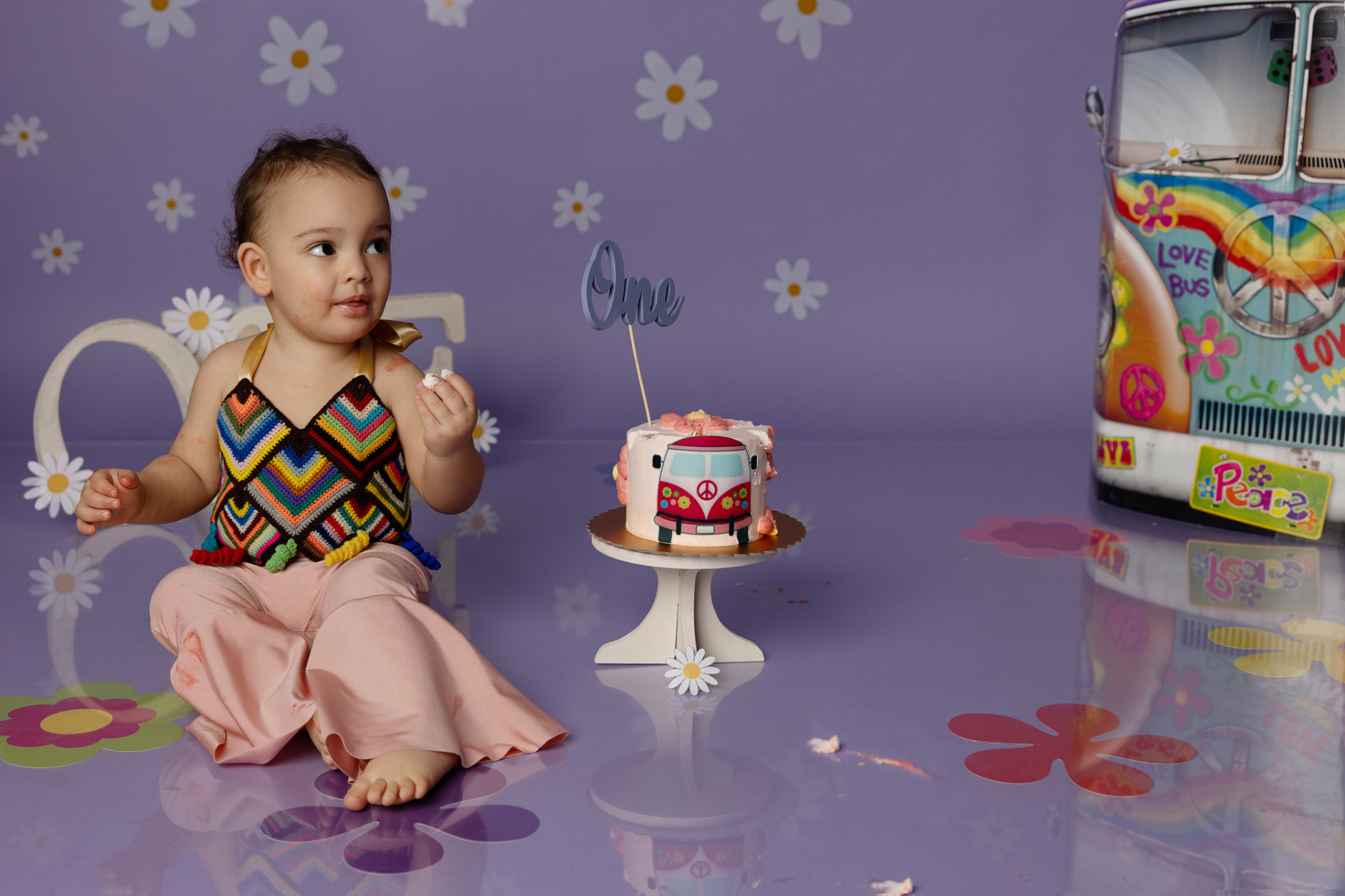 Bambina seduta accanto a torta di compleanno con topper "One", davanti alla lettera "O" e furgone decorativo. Pavimento viola con fiori e atmosfera anni '60. Foto di primo compleanno scattata da Claudia Spaziani nel suo studio a Bologna.