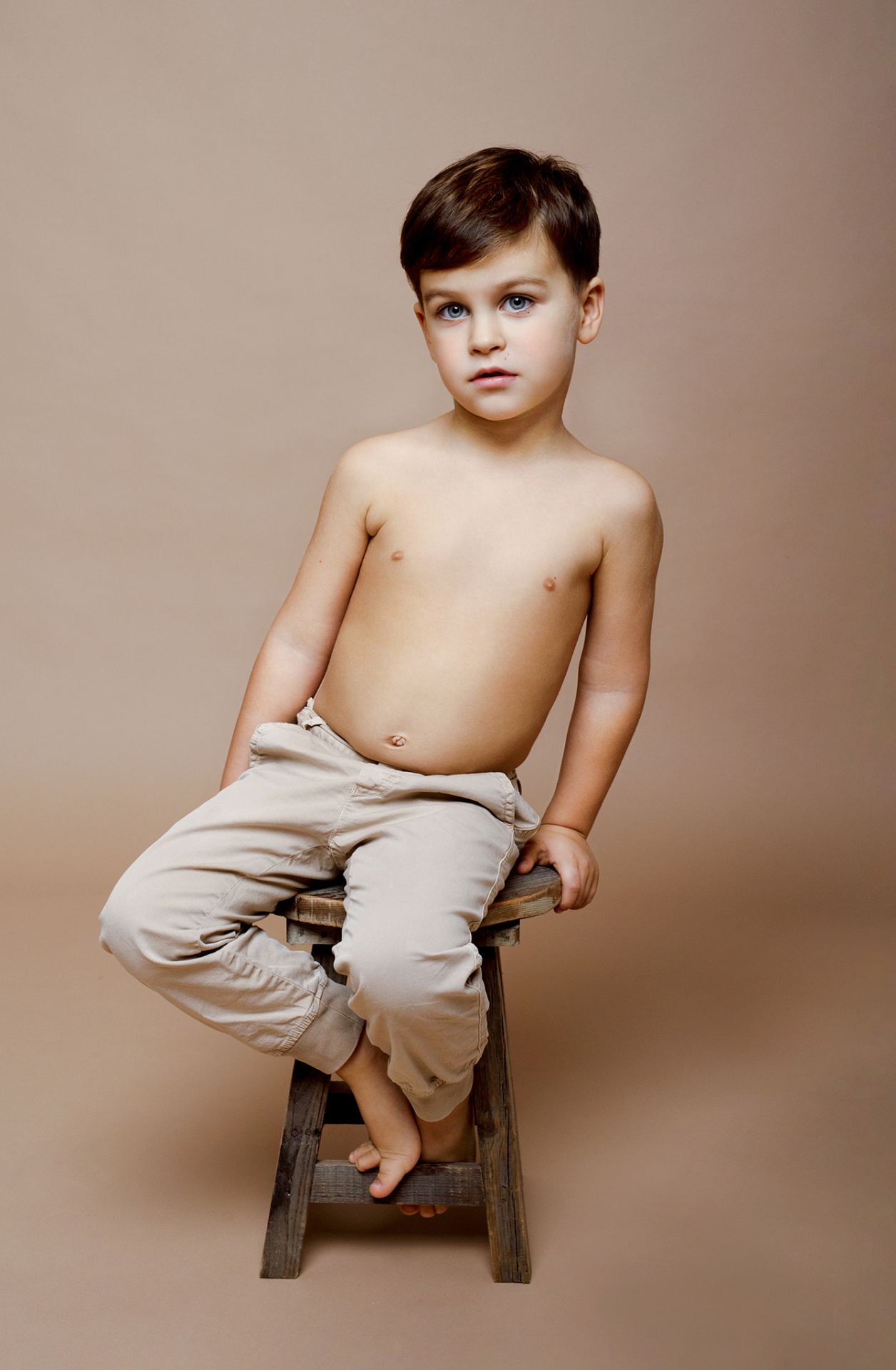 Ritratto semplice di bambino a torso nudo con pantaloni chiari, seduto su sgabello in legno, scattato da Claudia Spaziani nel suo studio fotografico a Bologna.