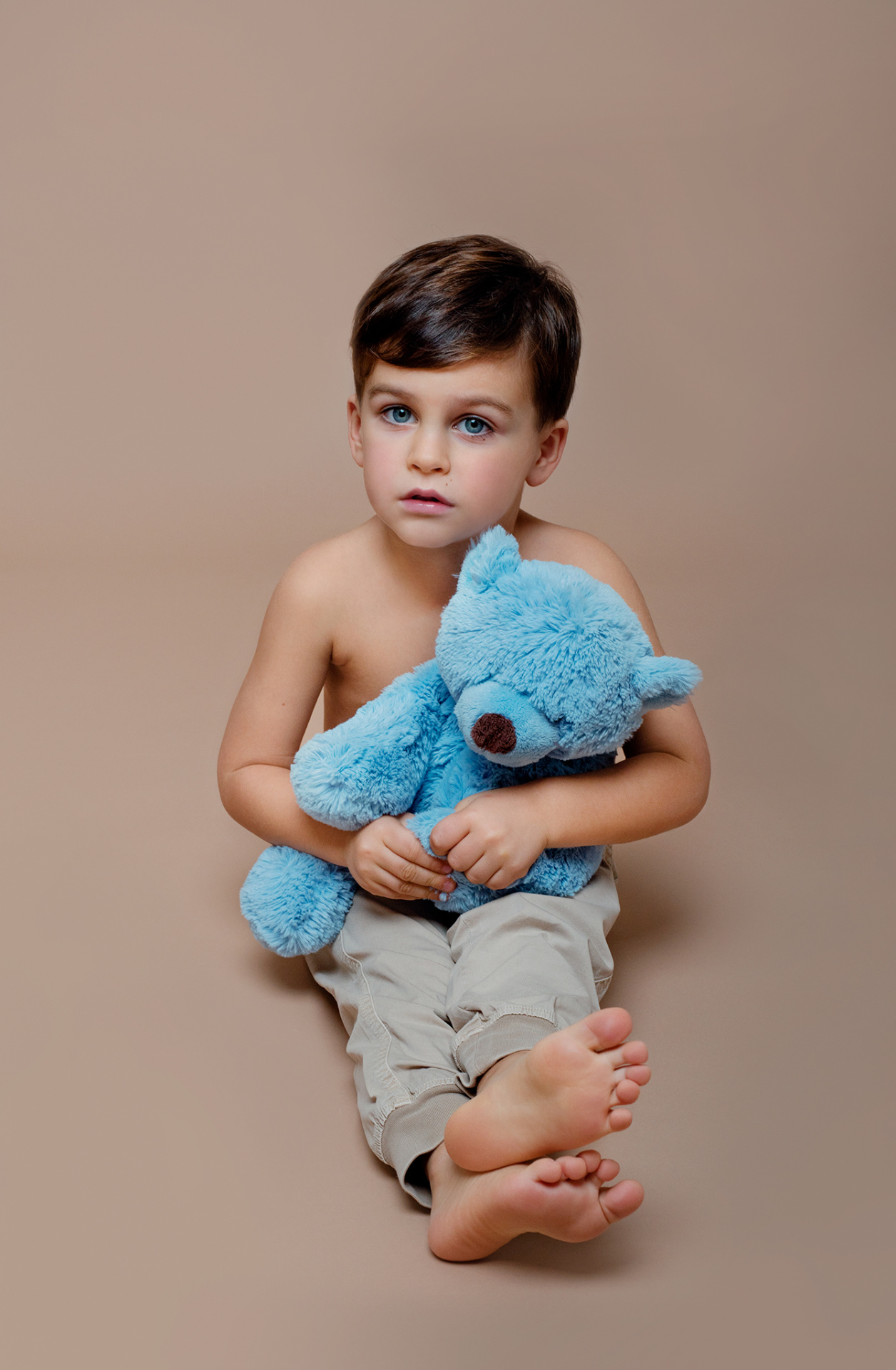 Ritratto tenero di bambino con orsetto blu, a piedi nudi su sfondo beige, realizzato da Claudia Spaziani nel suo studio fotografico a Bologna.