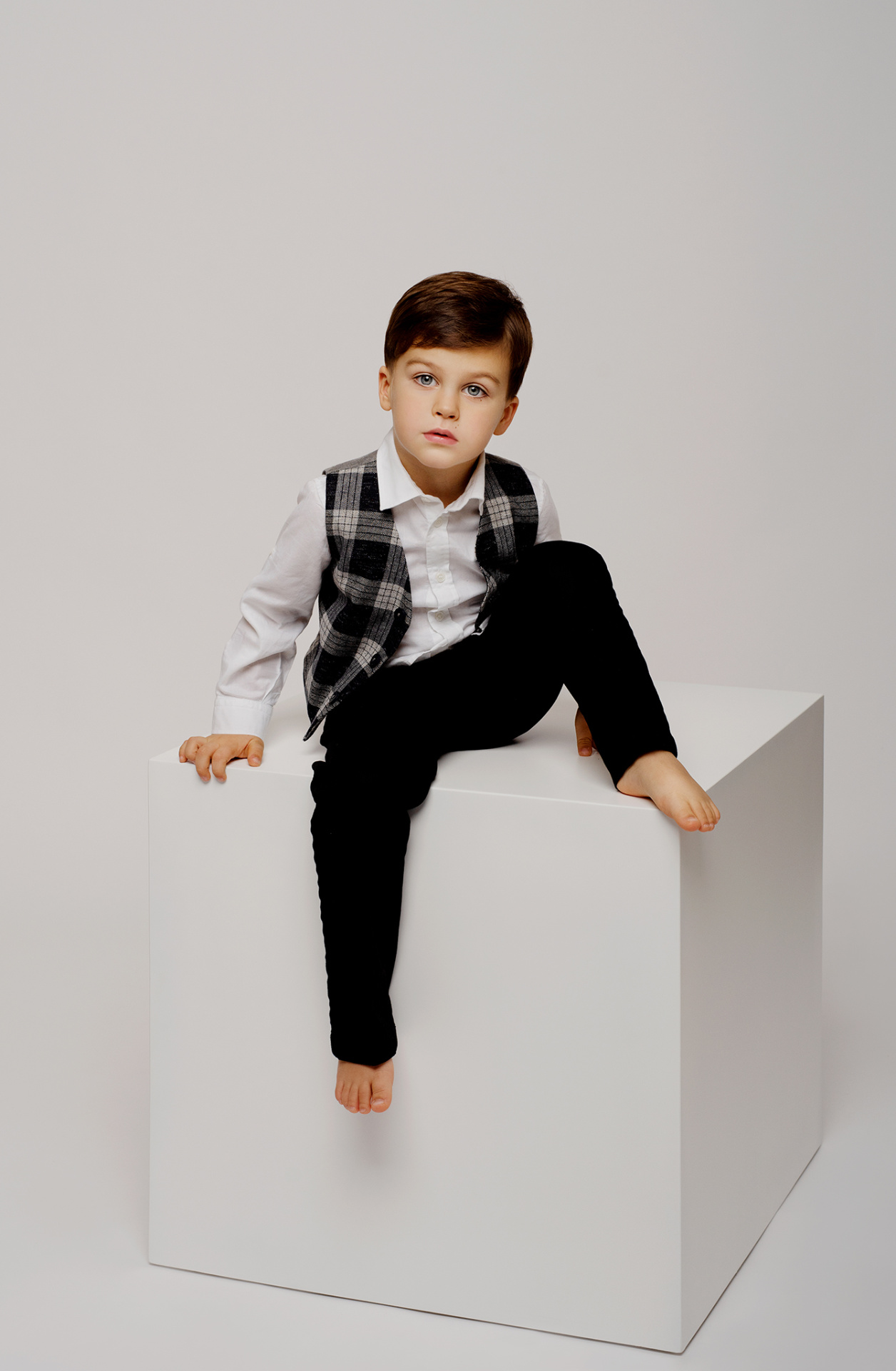 Ritratto minimalista di bambino in camicia bianca e gilet scozzese, seduto su cubo bianco, scattato da Claudia Spaziani nel suo studio fotografico a Bologna.