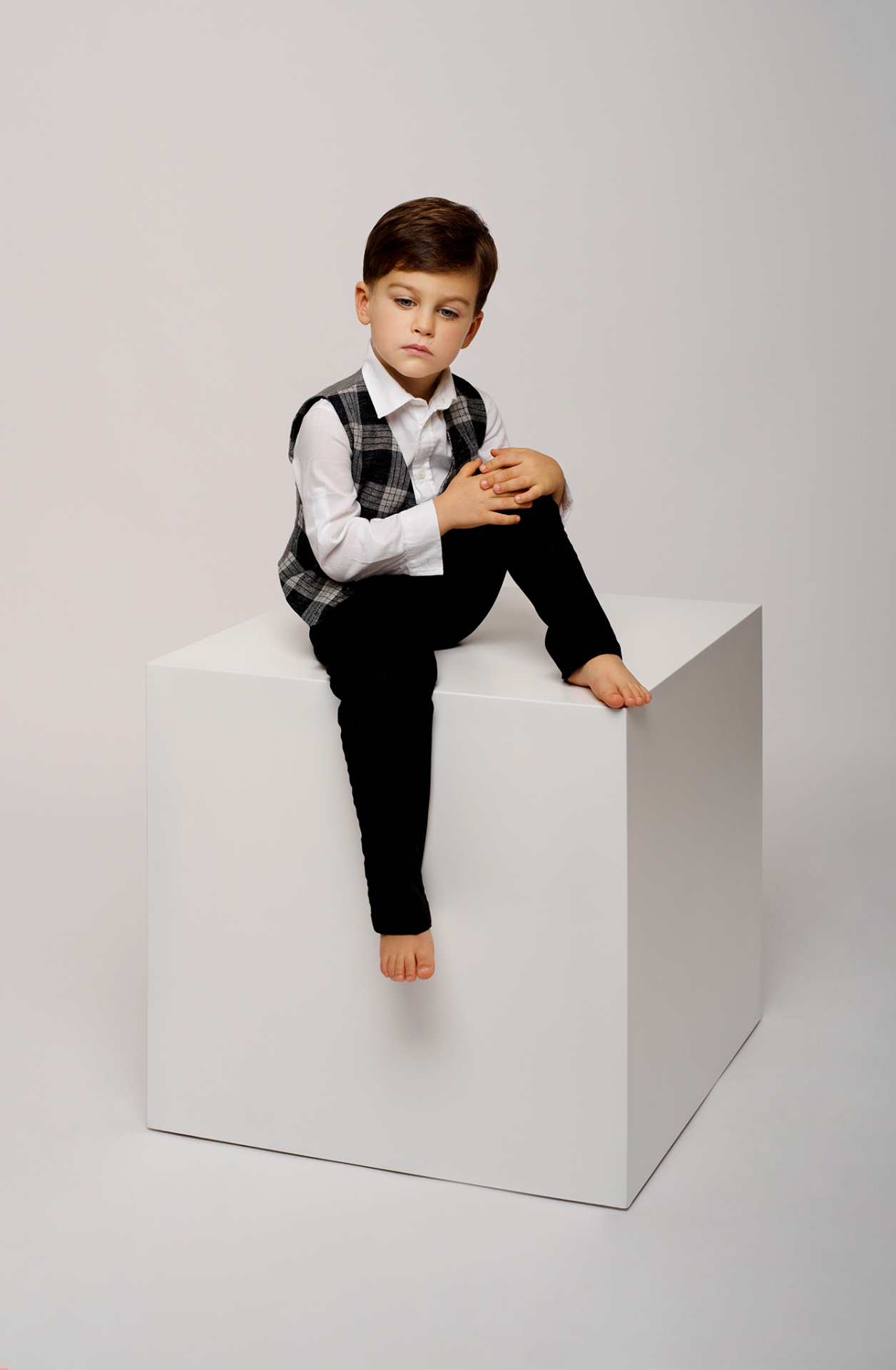 Ritratto in studio di bambino in camicia e gilet scozzese, seduto a piedi nudi su cubo bianco, realizzato da Claudia Spaziani a Bologna.
